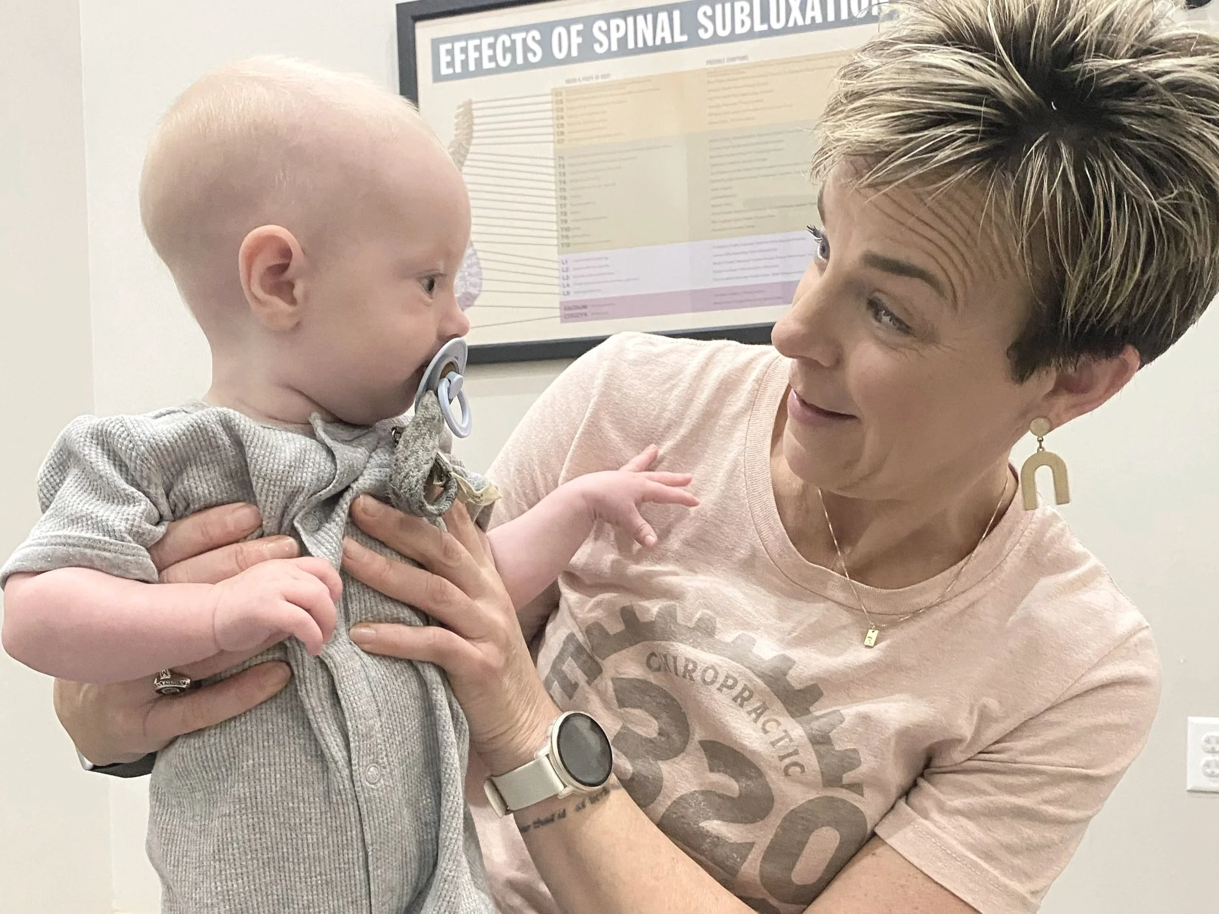 A woman holding a young child with a pacifier in his mouth, looking at each other. The woman has short, styled hair and is wearing a beige t-shirt. The child is wearing a gray, striped hoodie. In the background, there is a framed poster titled 'Effects of Spinal Subluxation' on the wall.