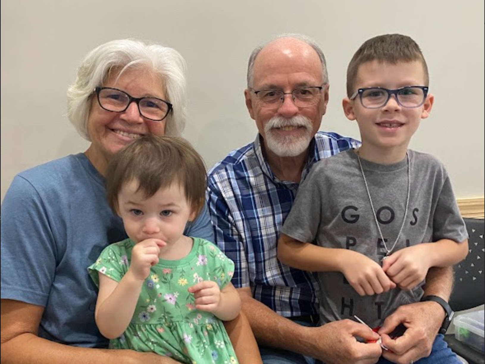 A group of five people, including two elderly adults and three children, posing together and smiling inside a room with plain walls.
