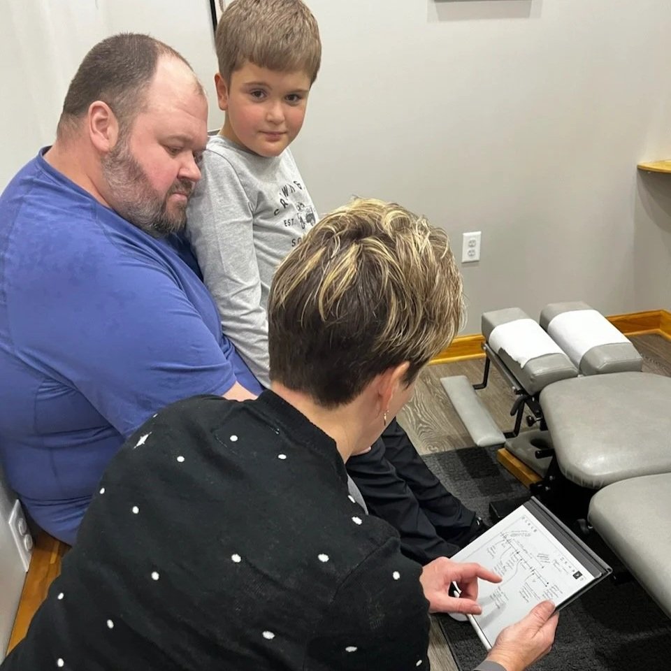 A woman shows a booklet to a man, a boy, and a girl in a waiting room with grey chairs.