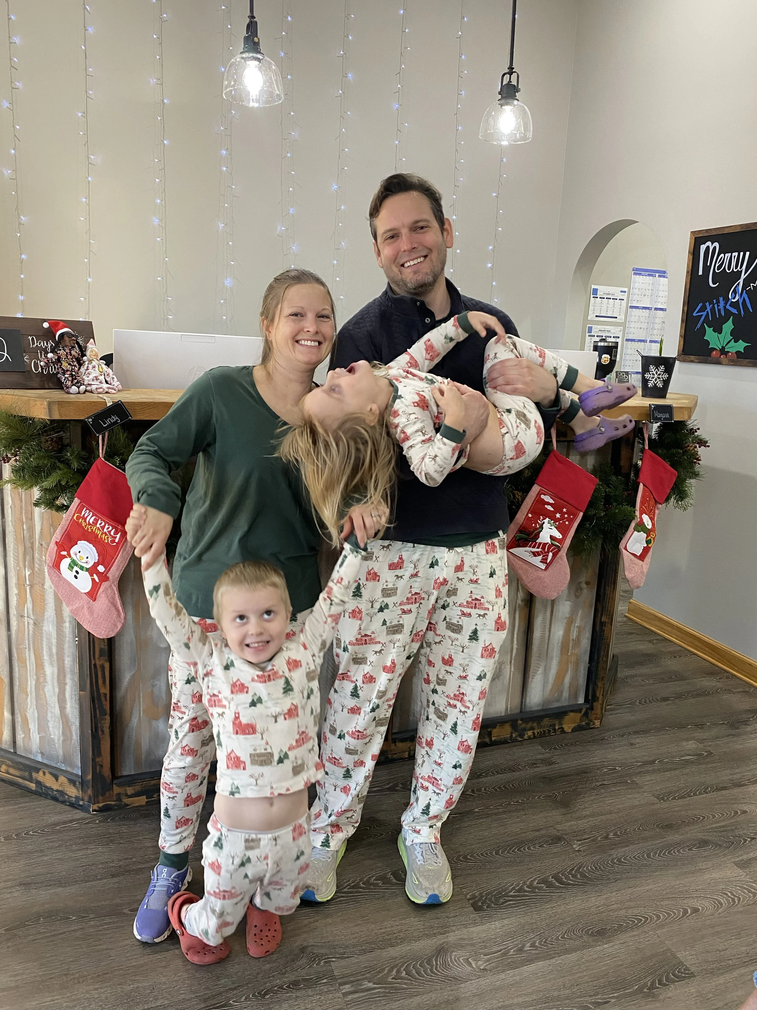 A man and a woman holding two children in Christmas pajamas in front of a decorated fireplace with stockings and Christmas decorations.