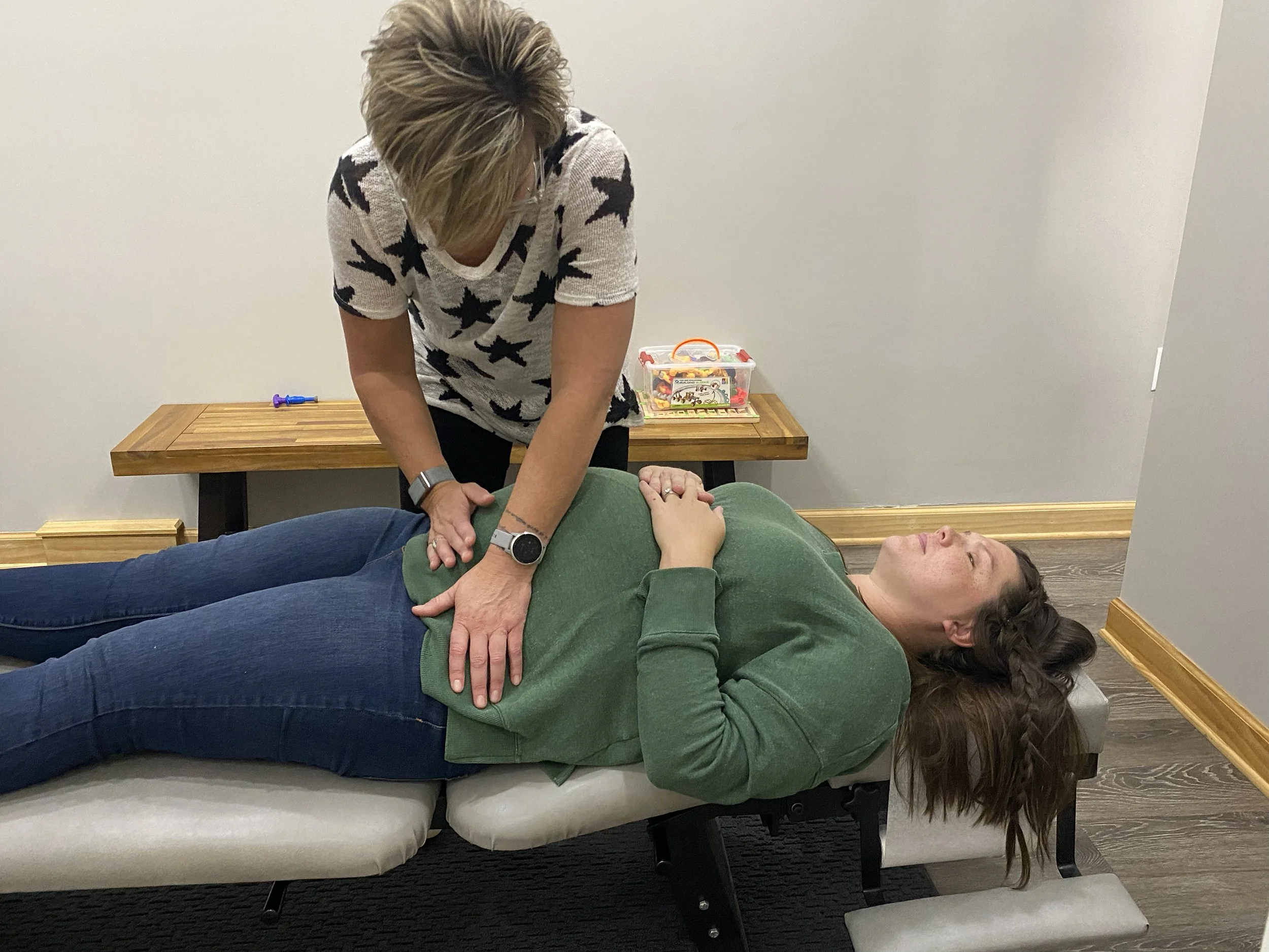 A woman performing CPR on a man lying on a chair in an indoor setting with wooden furniture and a plastic container in the background.
