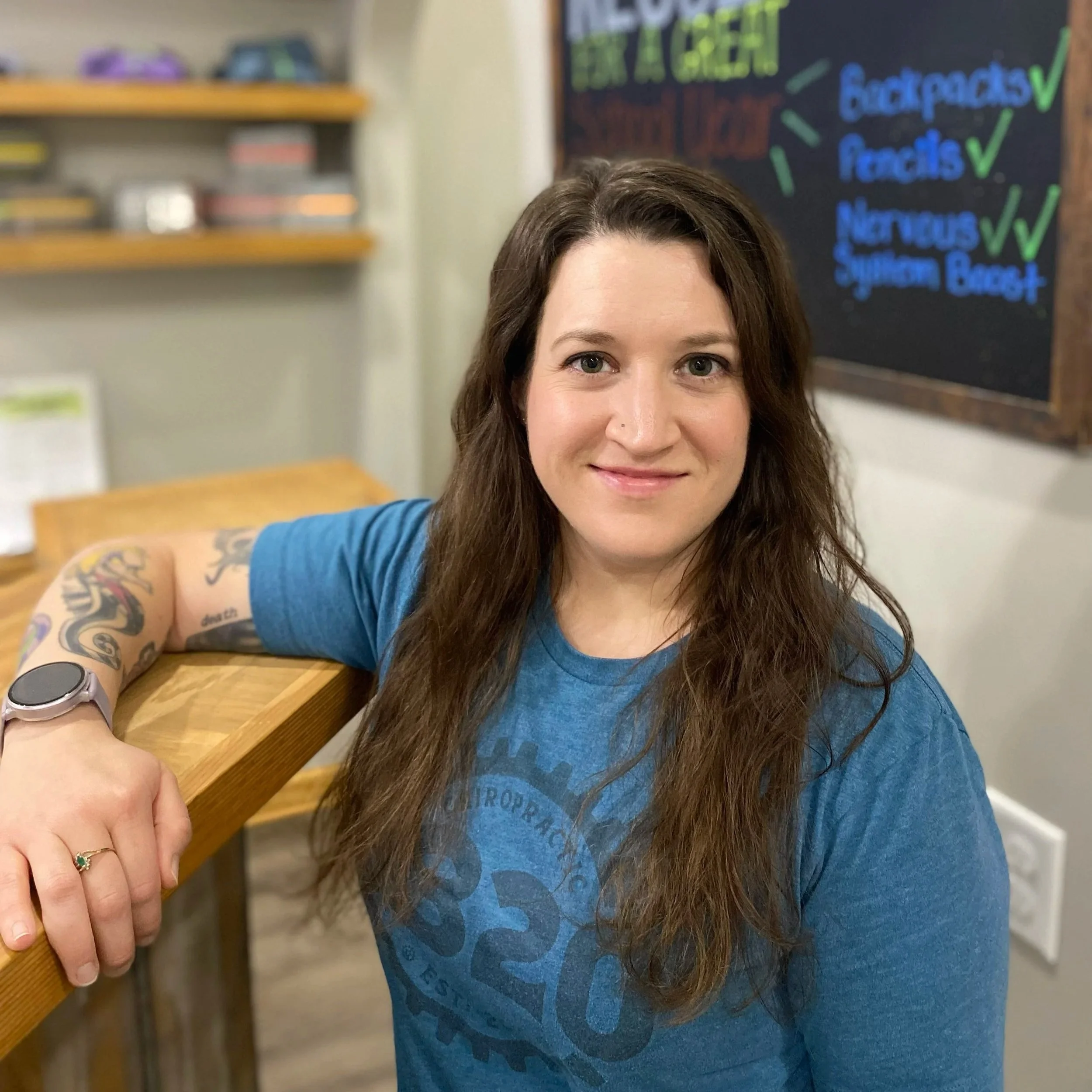 A woman with long brown hair and tattoos on her arm, wearing a blue T-shirt, is taking a selfie in a casual indoor setting. She is resting her arm on a wooden surface and has a ring with a green stone on her finger. In the background, there is a chalkboard with colorful writing and checkmarks, and shelves with various items.