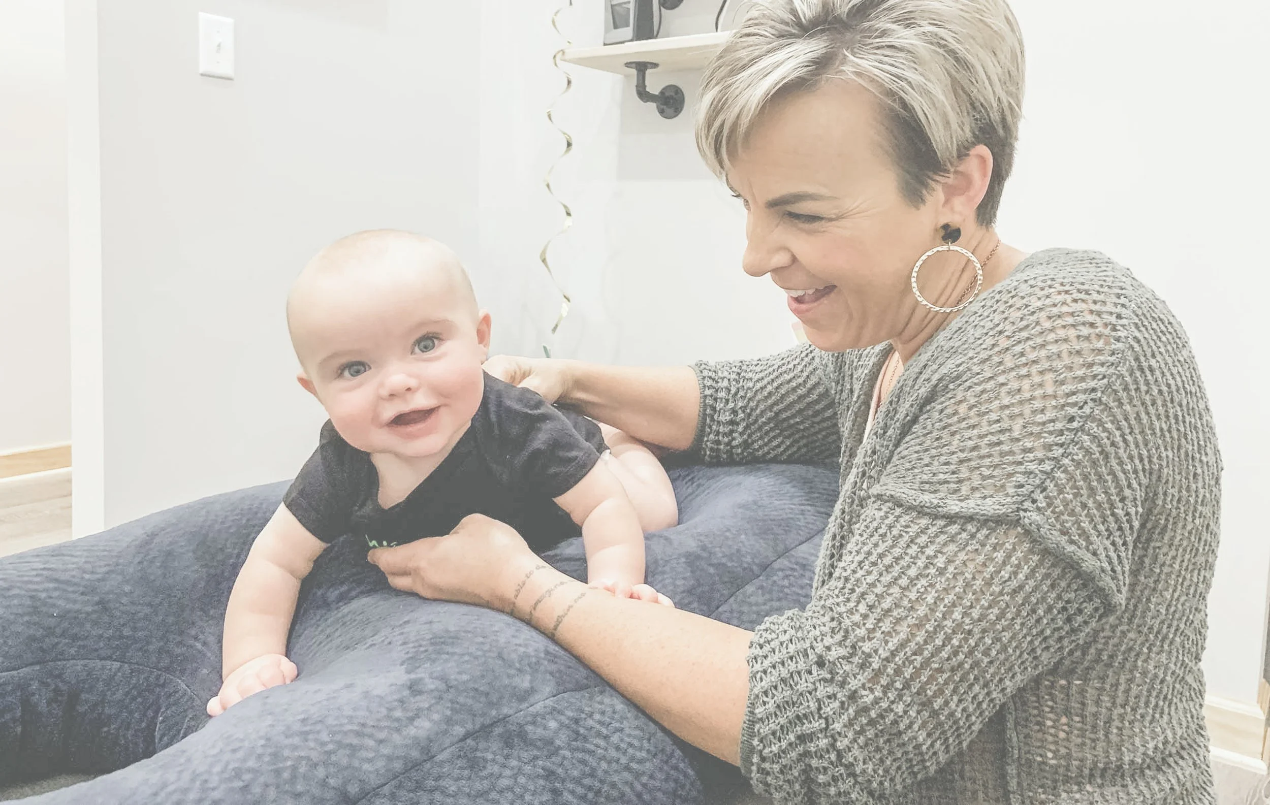 An older woman with short, light-colored hair and large hoop earrings is smiling and interacting with a smiling baby with light skin, blue eyes, and a shaved head, lying on a dark cushioned surface. The background includes a white wall, a shelf with decorative items, and some hanging streamers.