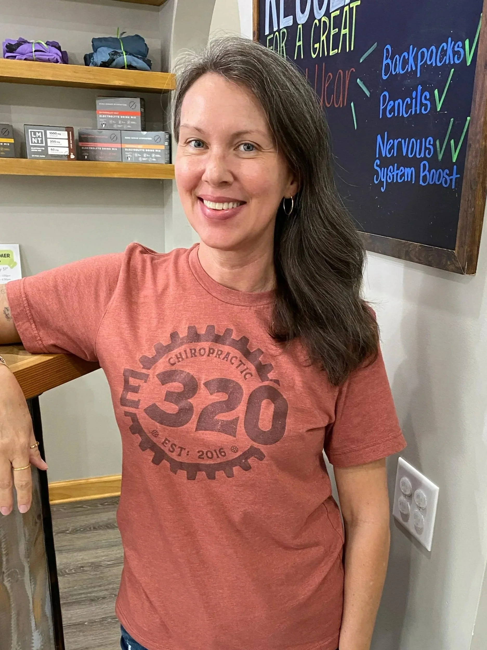 A woman with long brown hair and hoop earrings smiling at the camera, wearing a red T-shirt with a bicycle gear logo and text. Behind her, a blackboard with colorful writing and a shelf with folded clothes and boxes.