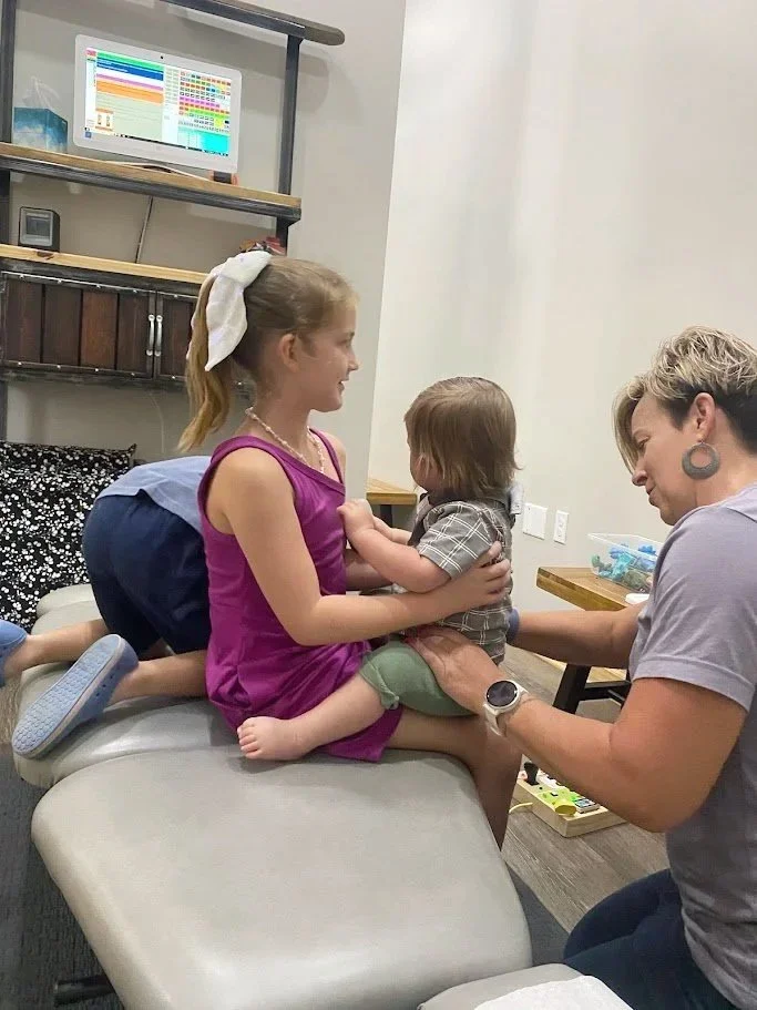 A young girl in a pink dress sitting on an examination table smiling at a small boy. The boy is sitting on her lap, facing her, and looking down. A woman, possibly a doctor or nurse, kneels beside them, helping or examining the boy. There is a computer monitor displaying a colorful schedule or chart on a desk in the background.