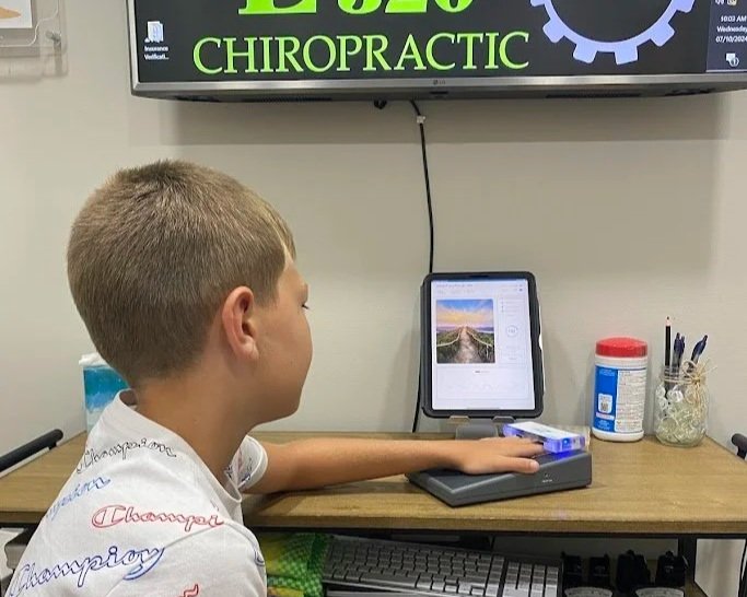 Young boy sitting at a desk watching a tablet, with a large computer monitor displayed above him showing the word 'Chiropractic,' and various office supplies on the desk.