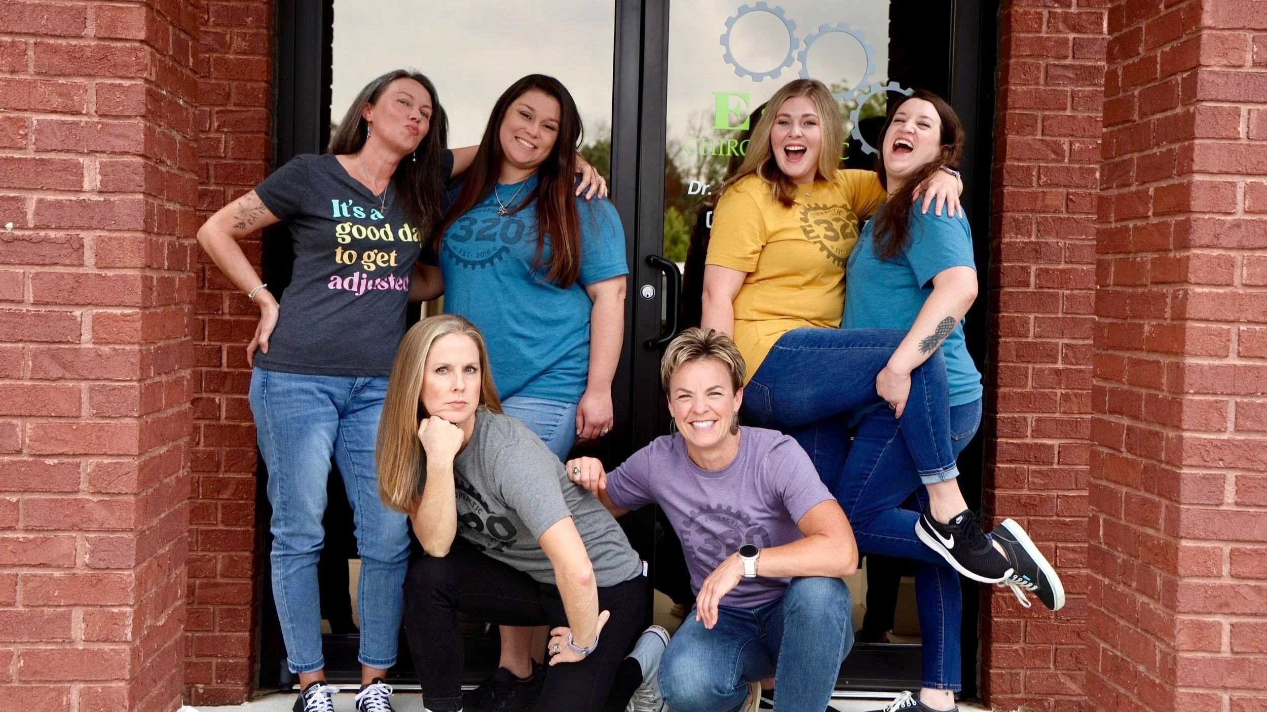 Group of women standing and sitting in front of a glass door with a brick wall on either side. They're smiling and showing camaraderie, wearing casual clothes, some with logos and text.