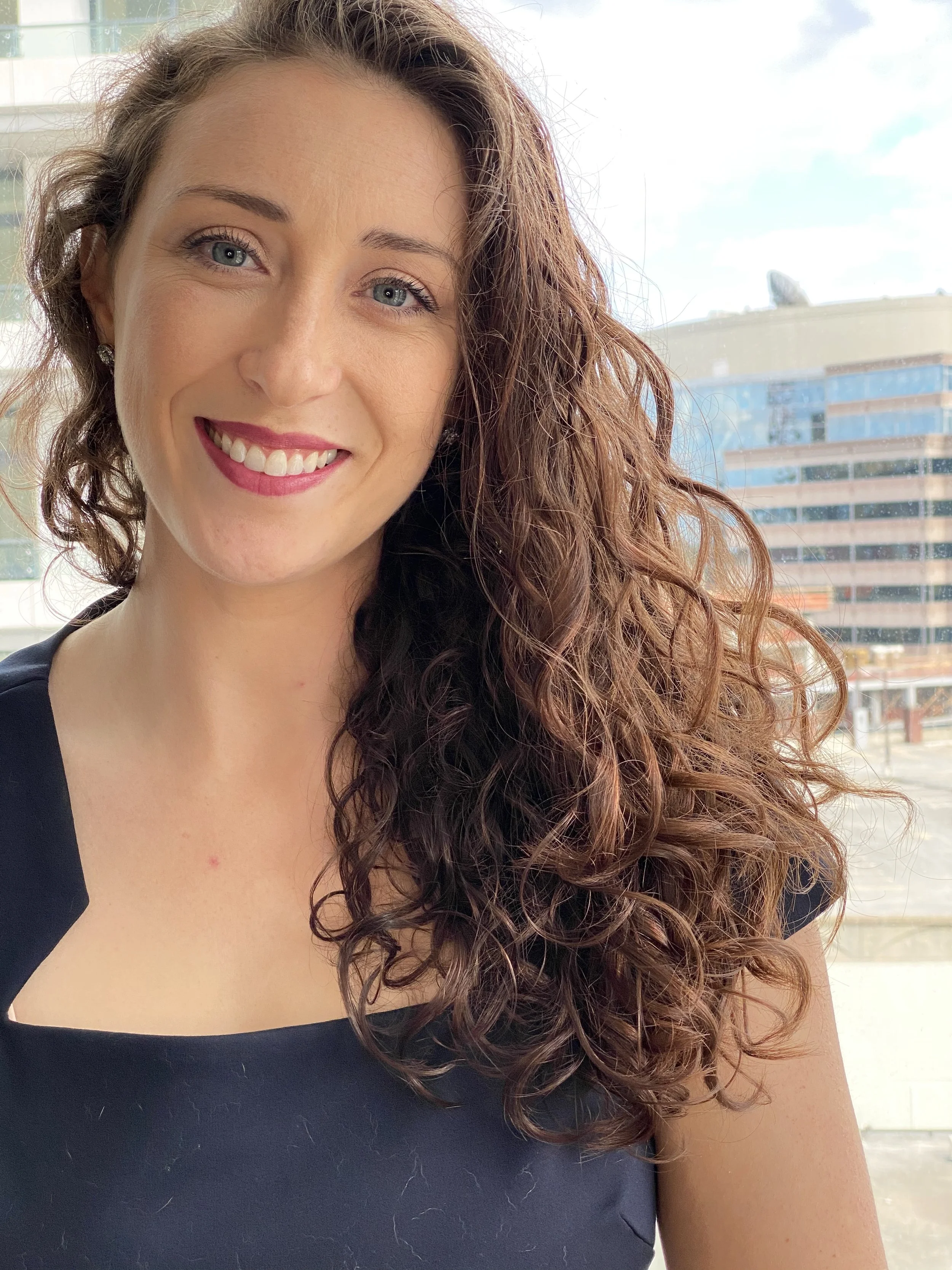 A smiling woman with curly auburn hair and blue eyes, wearing a black top, stands in front of a window with modern buildings outside.