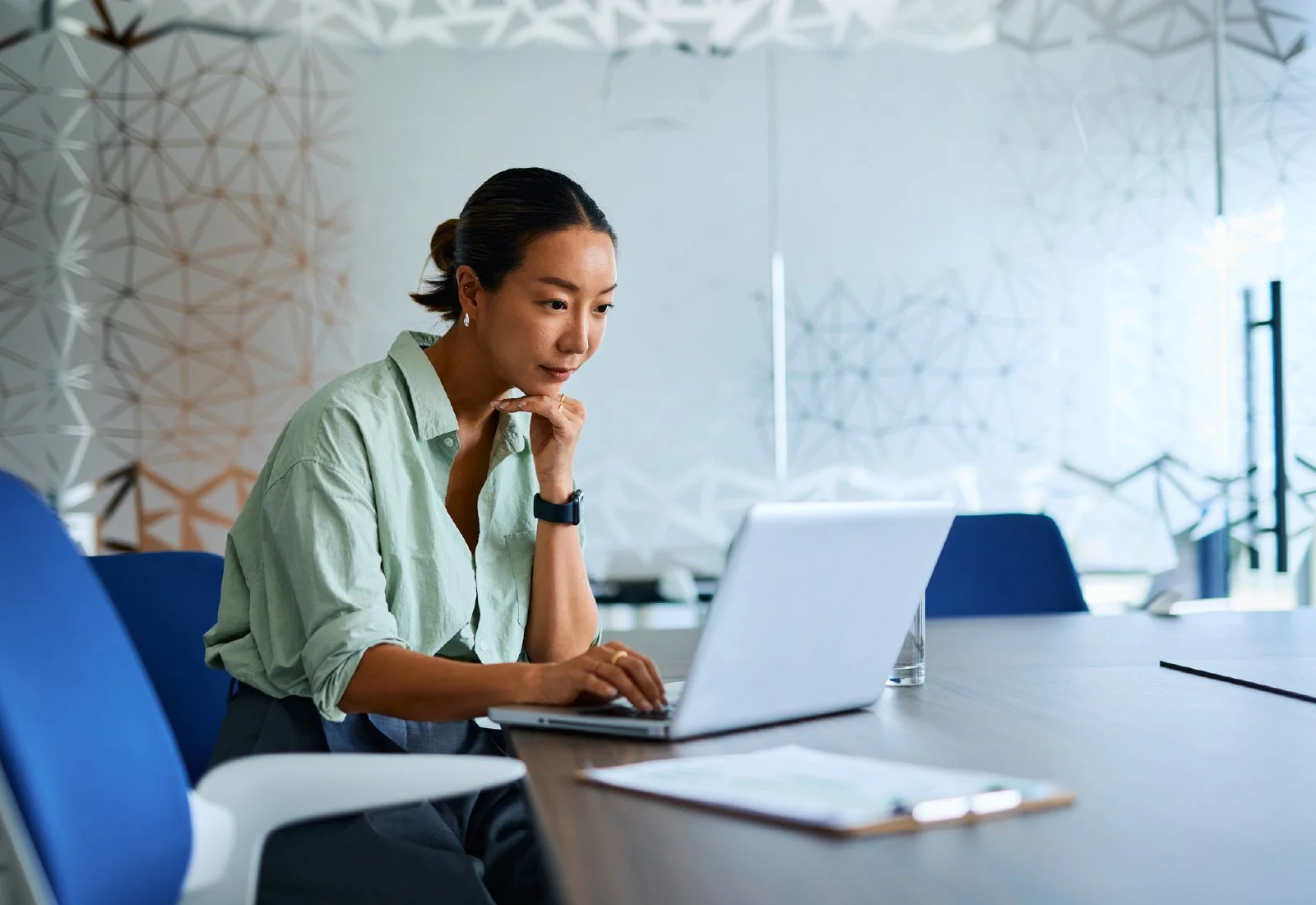 A woman sitting at a conference table working on her laptop in a modern office with geometric wall decorations.