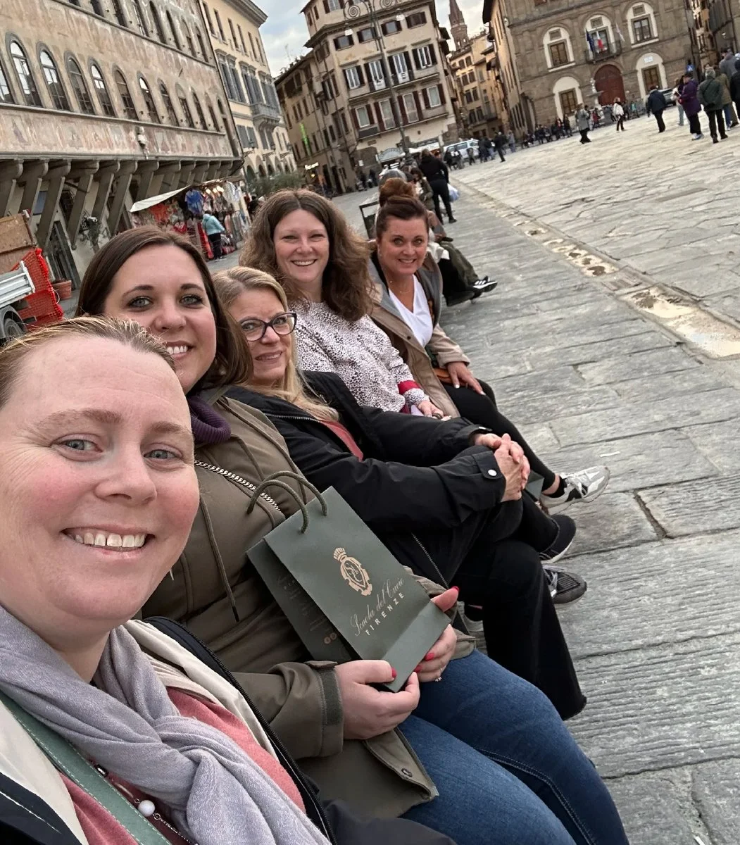 A group of five women sitting on a bench in a European city square, smiling at the camera, with historic buildings and people walking in the background.