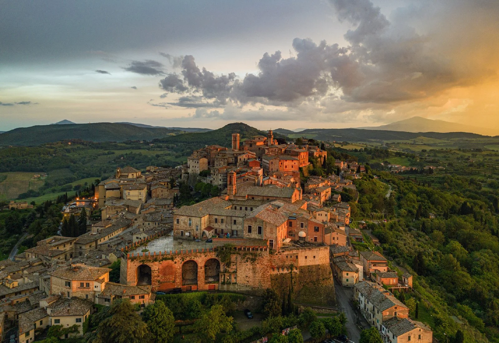 Aerial view of an ancient Italian hilltop town illuminated by sunset, with stone buildings, a fortress wall, and surrounded by green landscape and rolling hills.