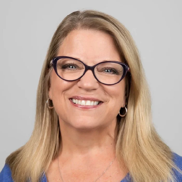 A woman with blonde hair wearing glasses and a blue top, smiling at the camera against a plain gray background.