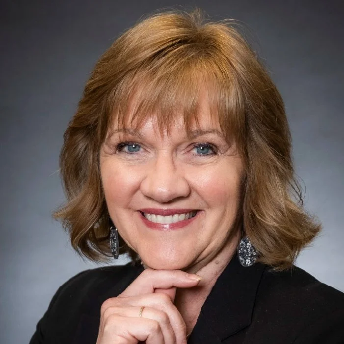 A woman with light brown hair, blue eyes, and a bright smile, wearing a black blazer and dangling earrings against a gray background.