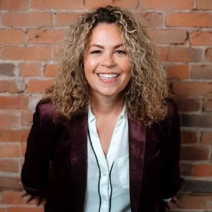 A smiling woman with curly blonde hair, wearing a white blouse and a dark blazer, standing in front of a brick wall.