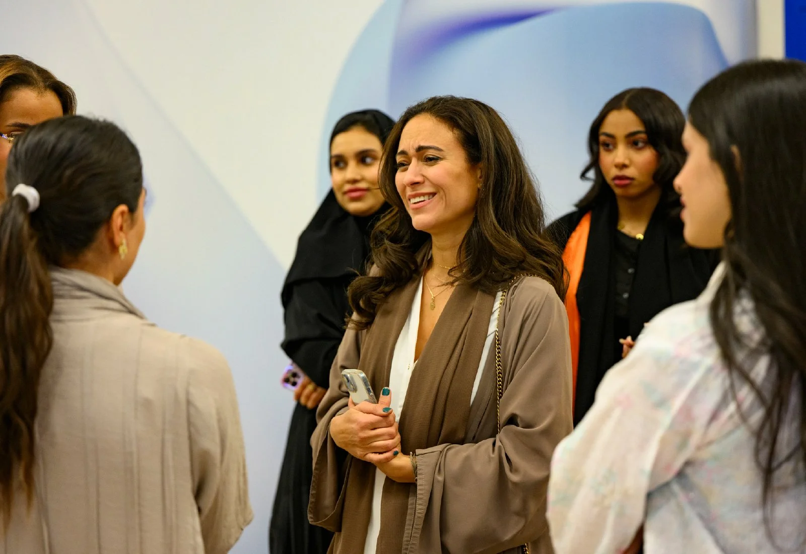 Group of women talking indoors, woman in brown cardigan smiling, holding a phone.