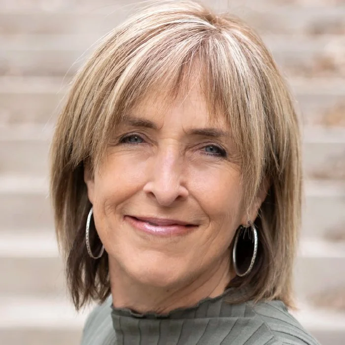 A woman with blonde hair smiling at the camera, wearing hoop earrings and a gray top, standing outdoors with steps in the background.