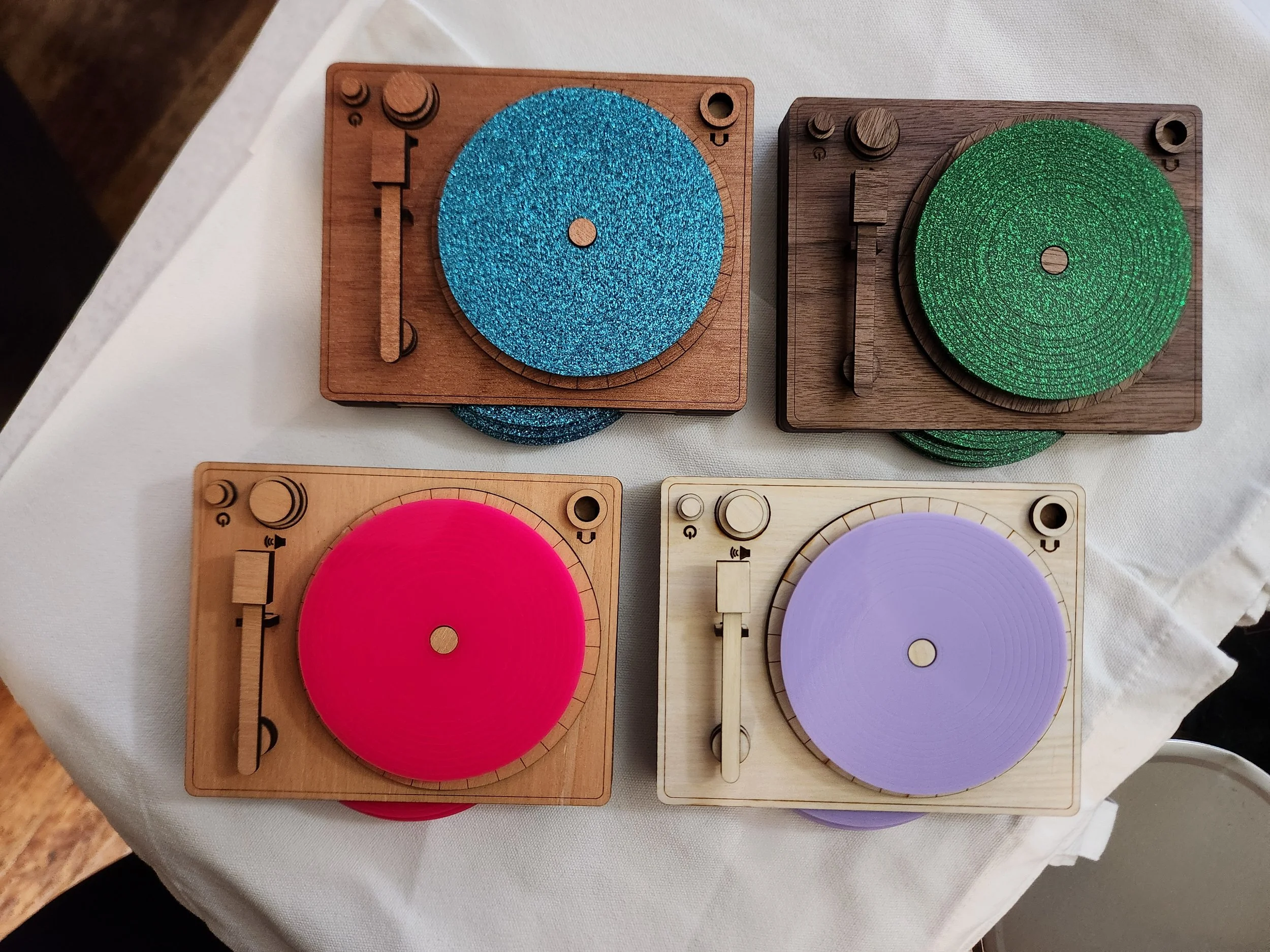 Four wooden turntables with colored accents, arranged on a white cloth. The top left turntable has a blue glittery platter, the top right has a green glittery platter, the bottom left has a bright pink platter, and the bottom right has a light purple platter.