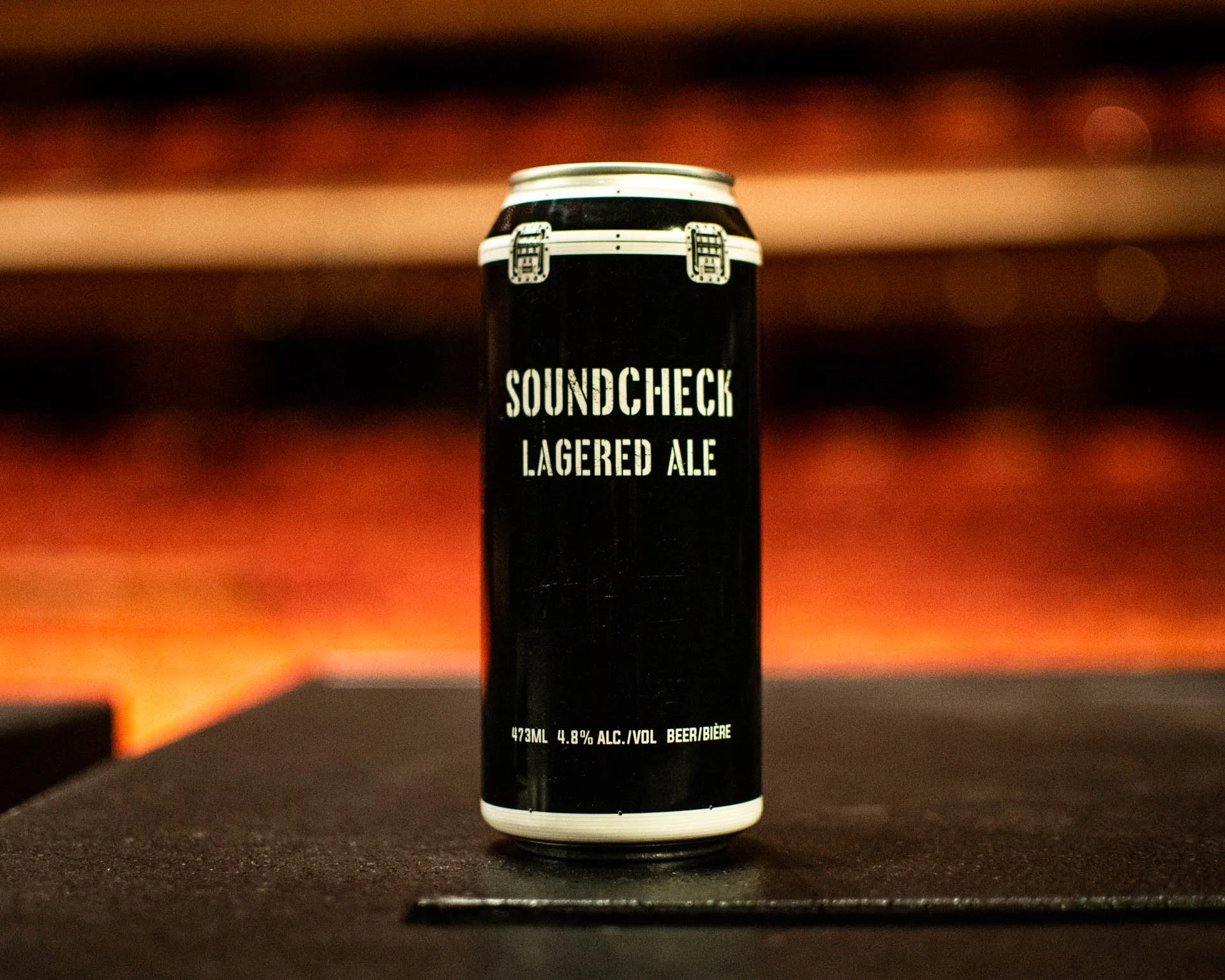 A black can of Soundcheck Lagered Ale with white text, standing on a dark surface against a blurred reddish-orange background.