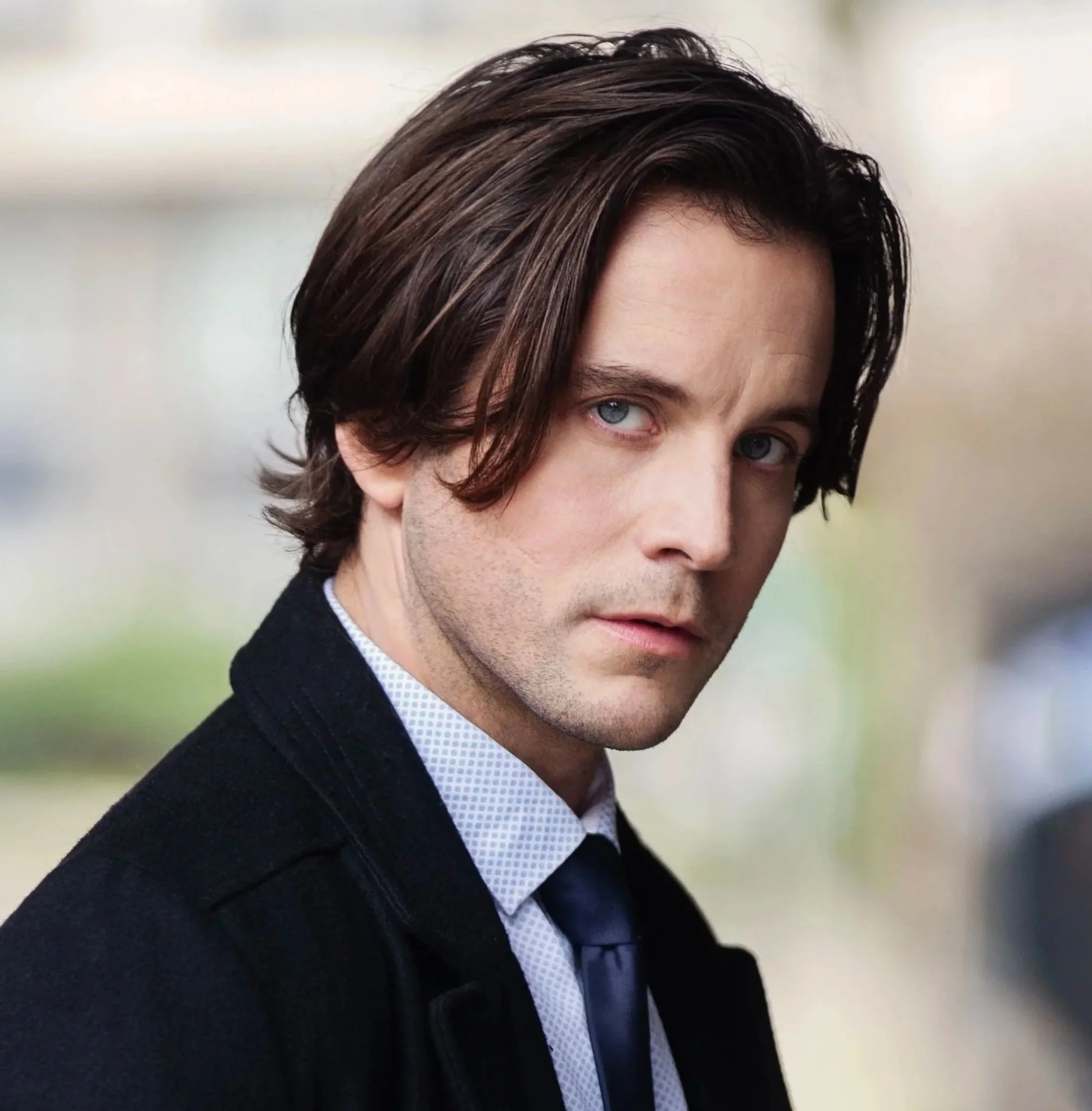 Close-up portrait of a young man with blue eyes, dark brown hair, and light facial hair, wearing a black shirt against a dark background.