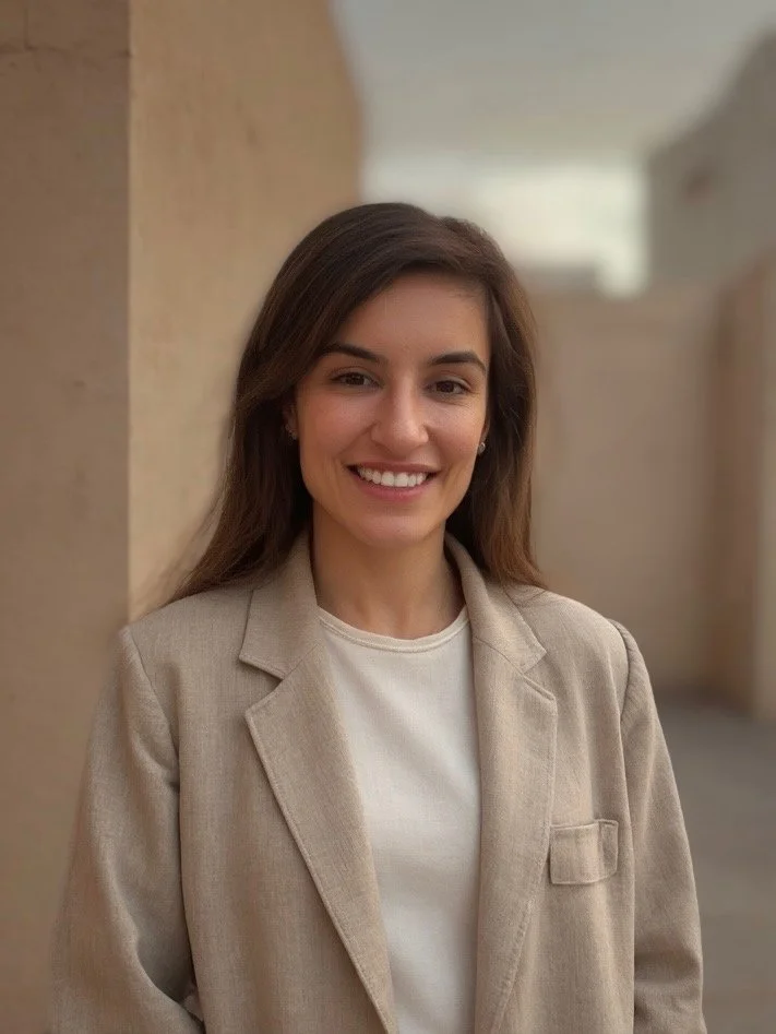A young woman with long brown hair, smiling, wearing a beige blazer and a white shirt, standing outdoors against a blurred beige wall background.