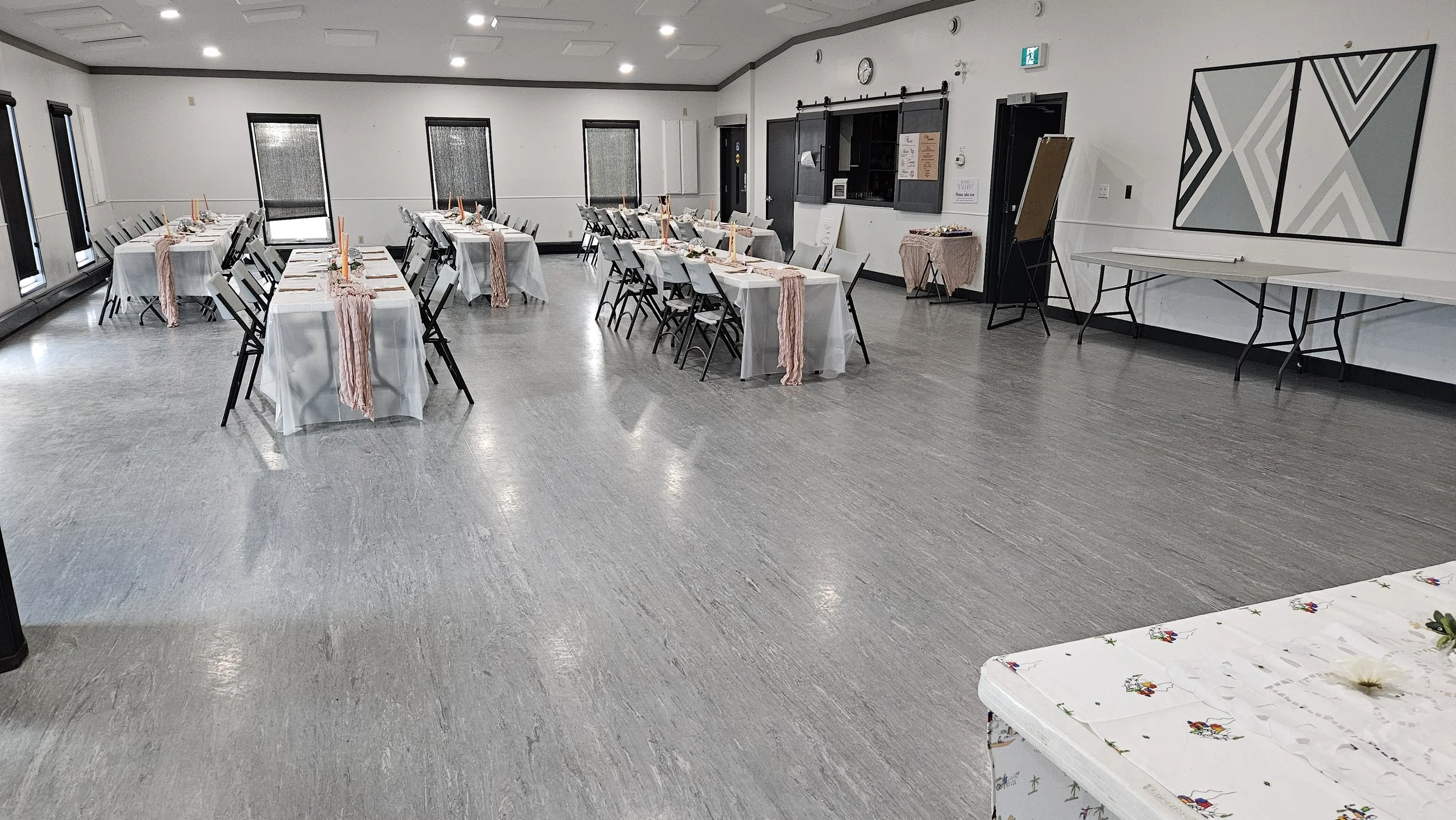 Large empty event hall with decorated tables and chairs, white tablecloths, and pink runners, prepared for a function or celebration.