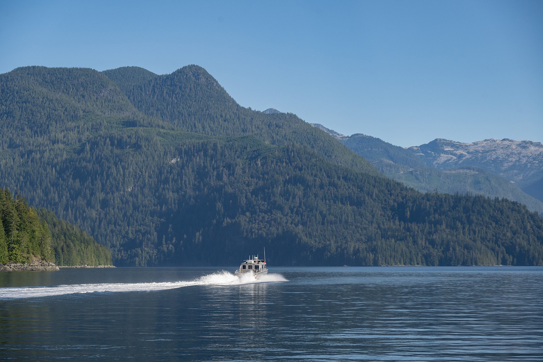 a boat driving away into the distance along blue inland waters, with forested mountains in the background