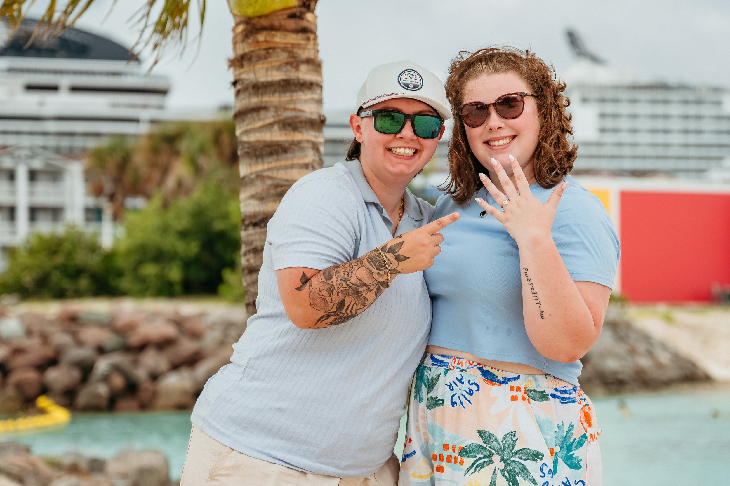 Two women smiling and posing for a photo outdoors, one showing off a ring on her hand, with a palm tree and a cruise ship in the background.