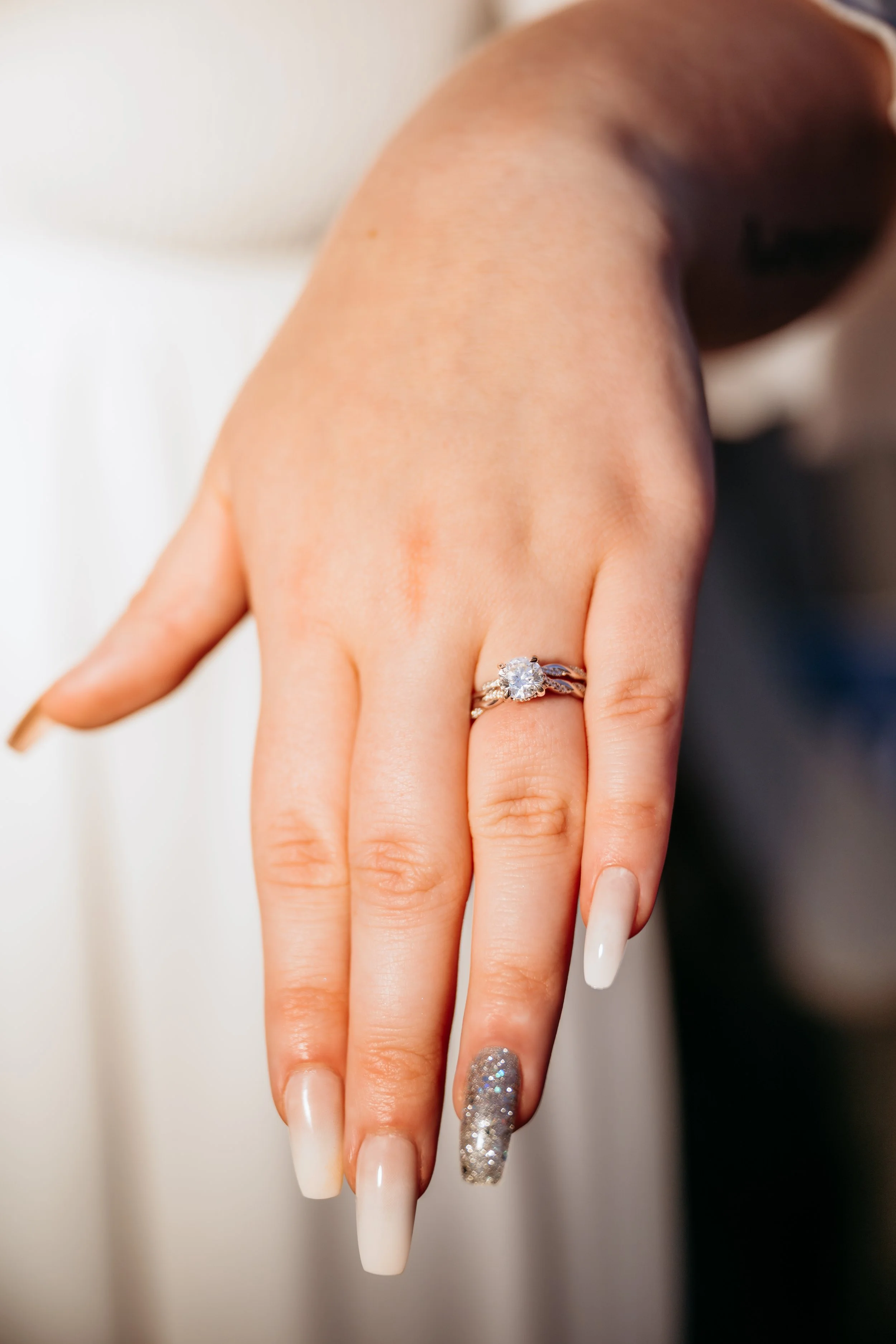Close-up of a woman's left hand displaying an engagement ring with a large central diamond and a wedding band, with manicure featuring nude and sparkly silver nails.