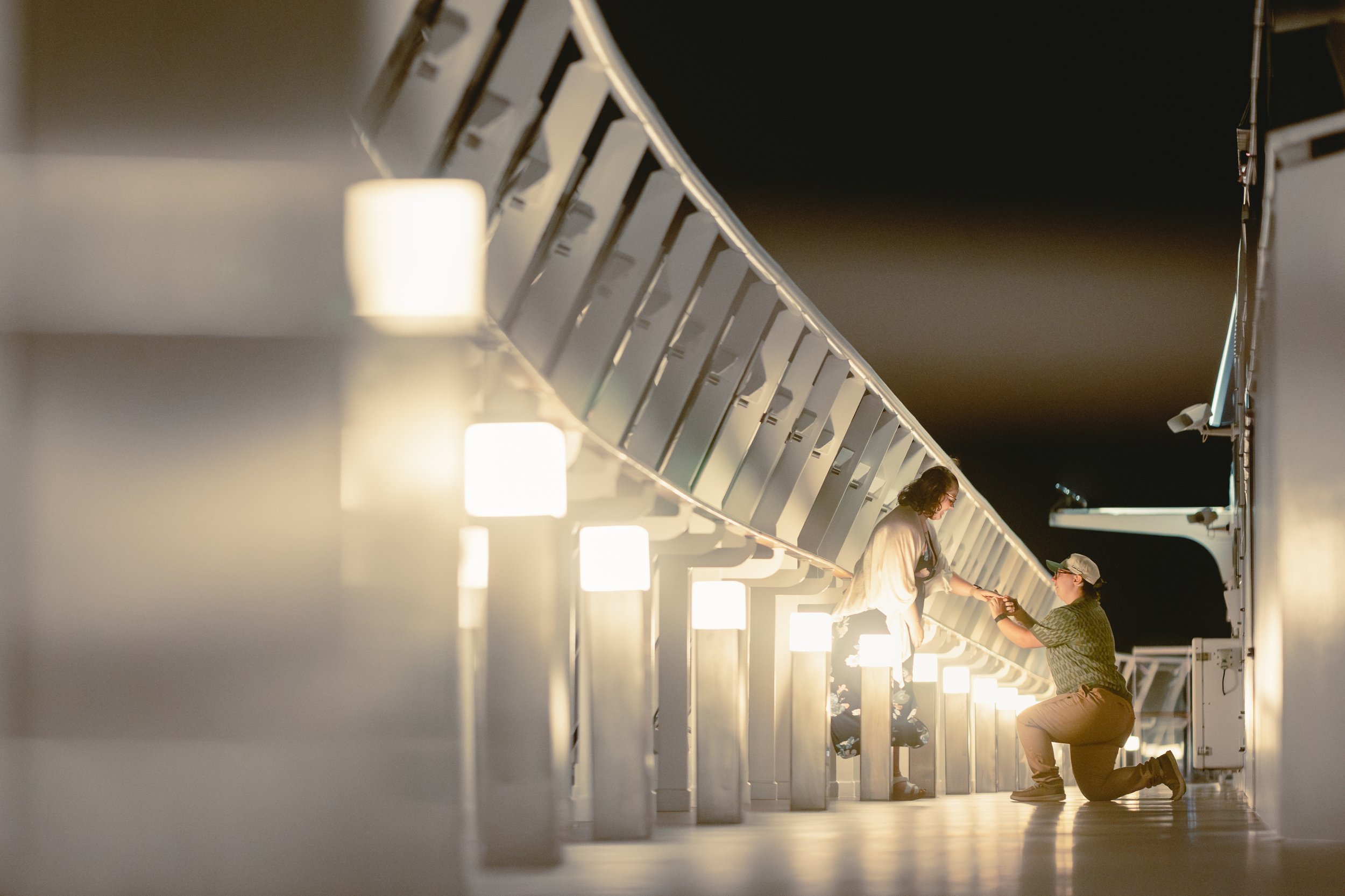 A man is kneeling on one knee on a ship deck at night, proposing to a woman who is standing on the deck. The scene is illuminated by a row of lights along the ship's railing.