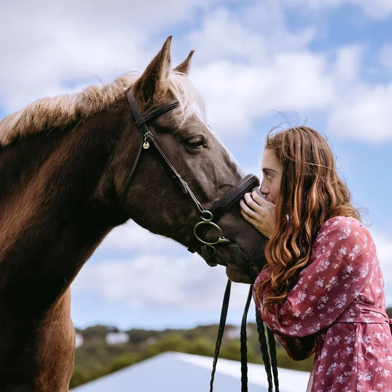 A girl with long red hair wearing a pink floral dress gently touching noses with a brown horse against a blue sky with clouds.