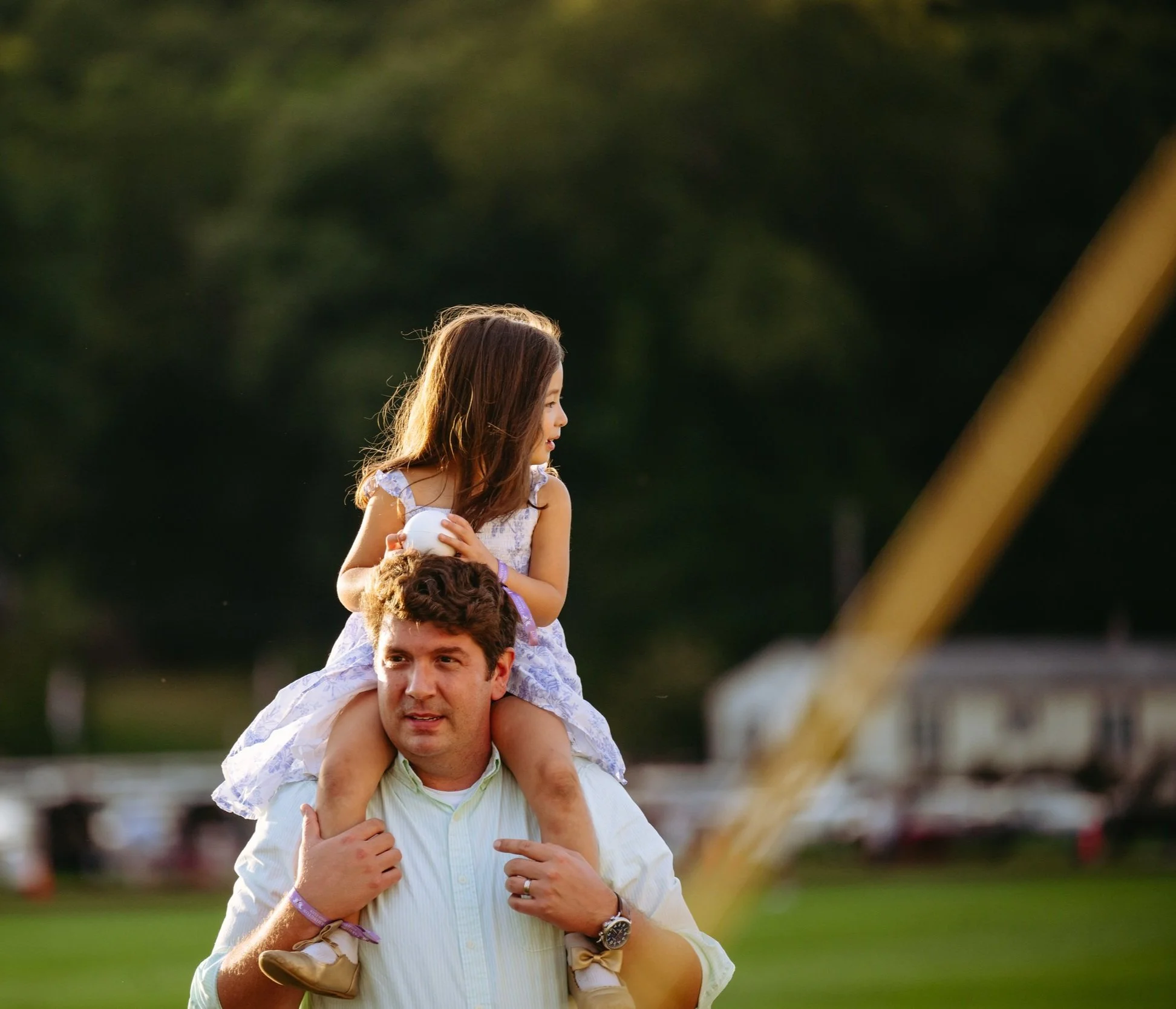 A man carrying a young girl on his shoulders in an outdoor setting during daylight, with a blurred background of trees and open space.
