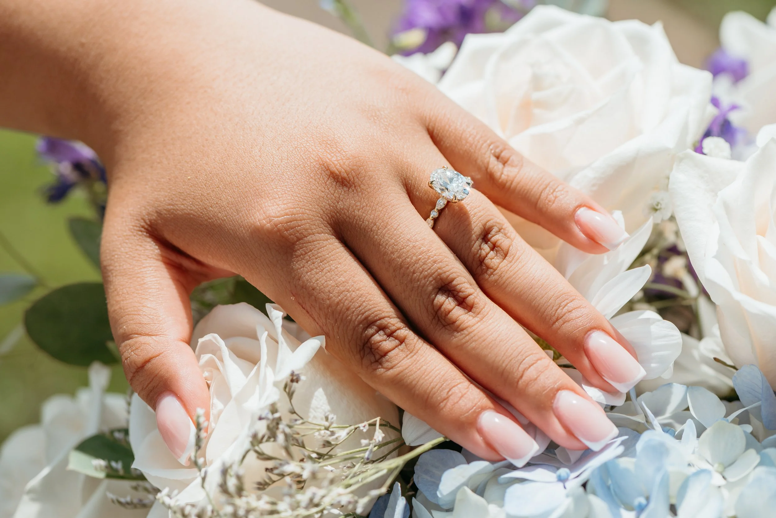A hand with an engagement ring rests on a bed of white and purple flowers.