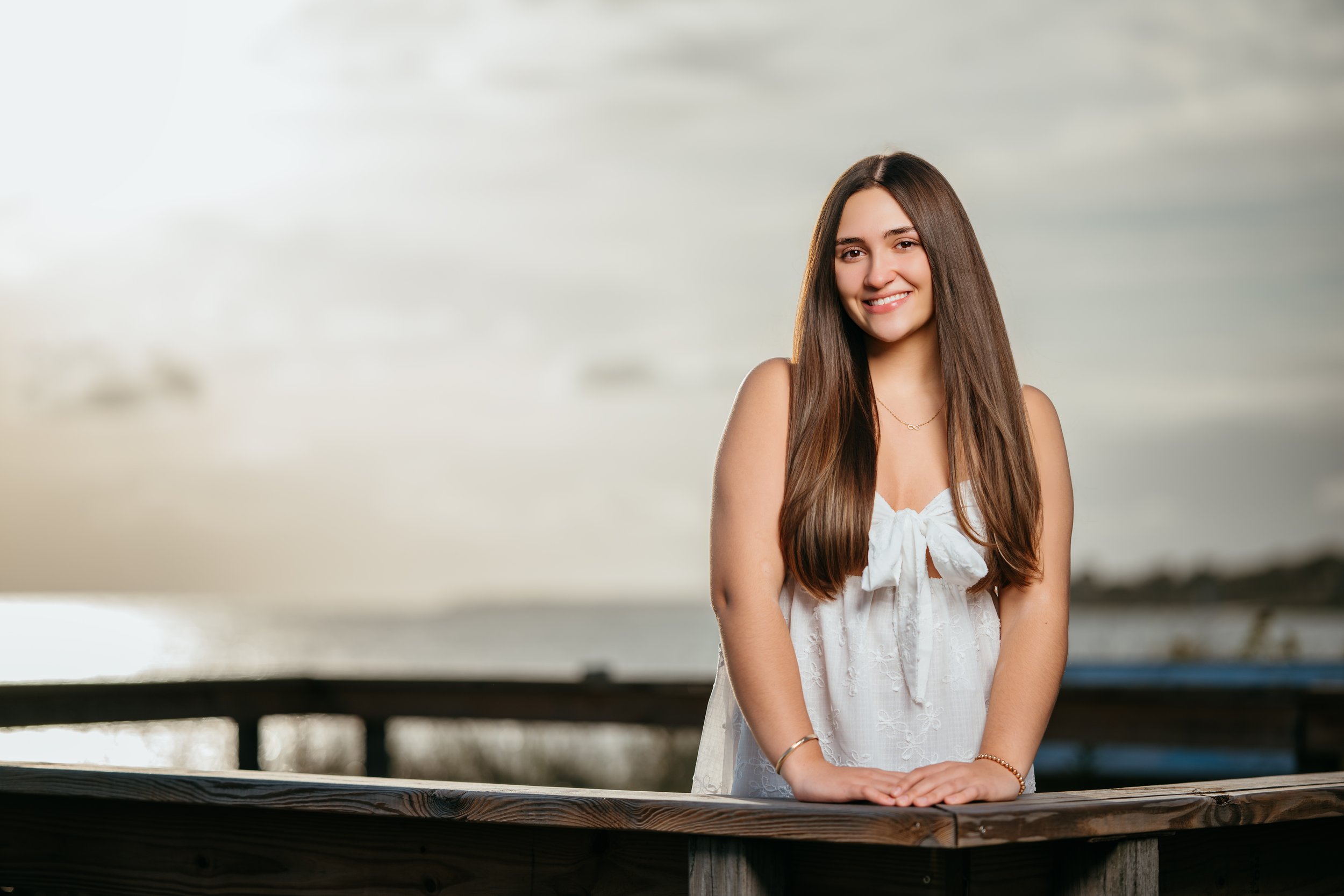 A young woman with long brown hair smiling, wearing a white sleeveless top with a front bow, standing outdoors by a wooden railing near water, with an overcast sky in the background.