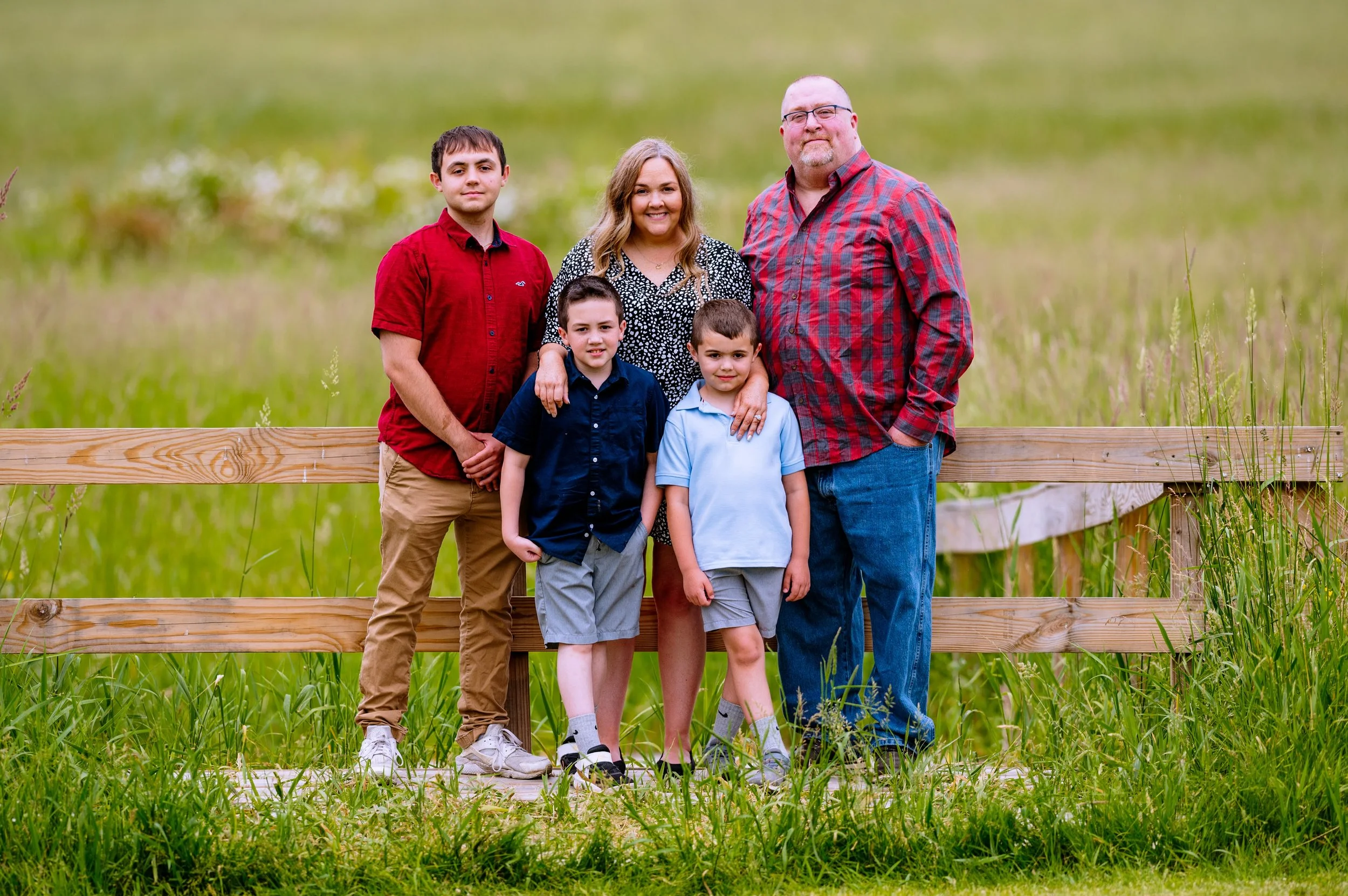 Family of five posing outdoors in front of a wooden fence, with green grass and a blurred field background. Two adults and three children, all smiling at the camera.
