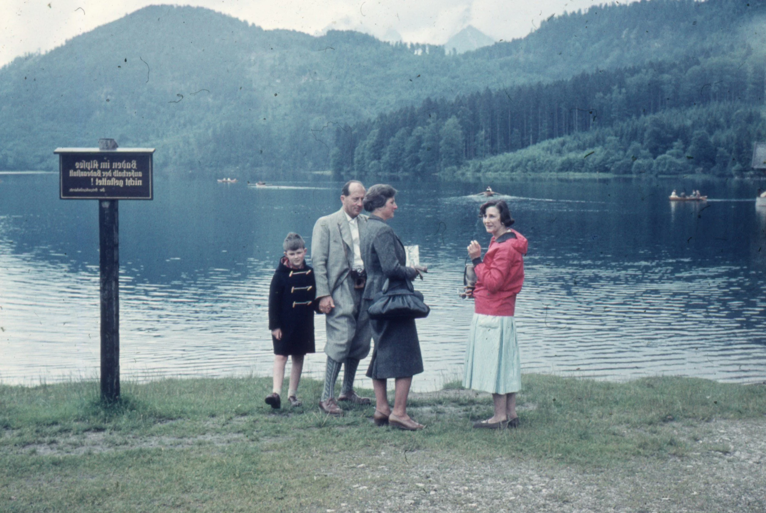 A group of five people, three women and two young boys, standing near a lake with mountains in the background. Two women are engaged in conversation, one with a red jacket and the other with a darker coat. The other woman and the boys are looking on. There are small boats on the lake and a sign on a post near the water.