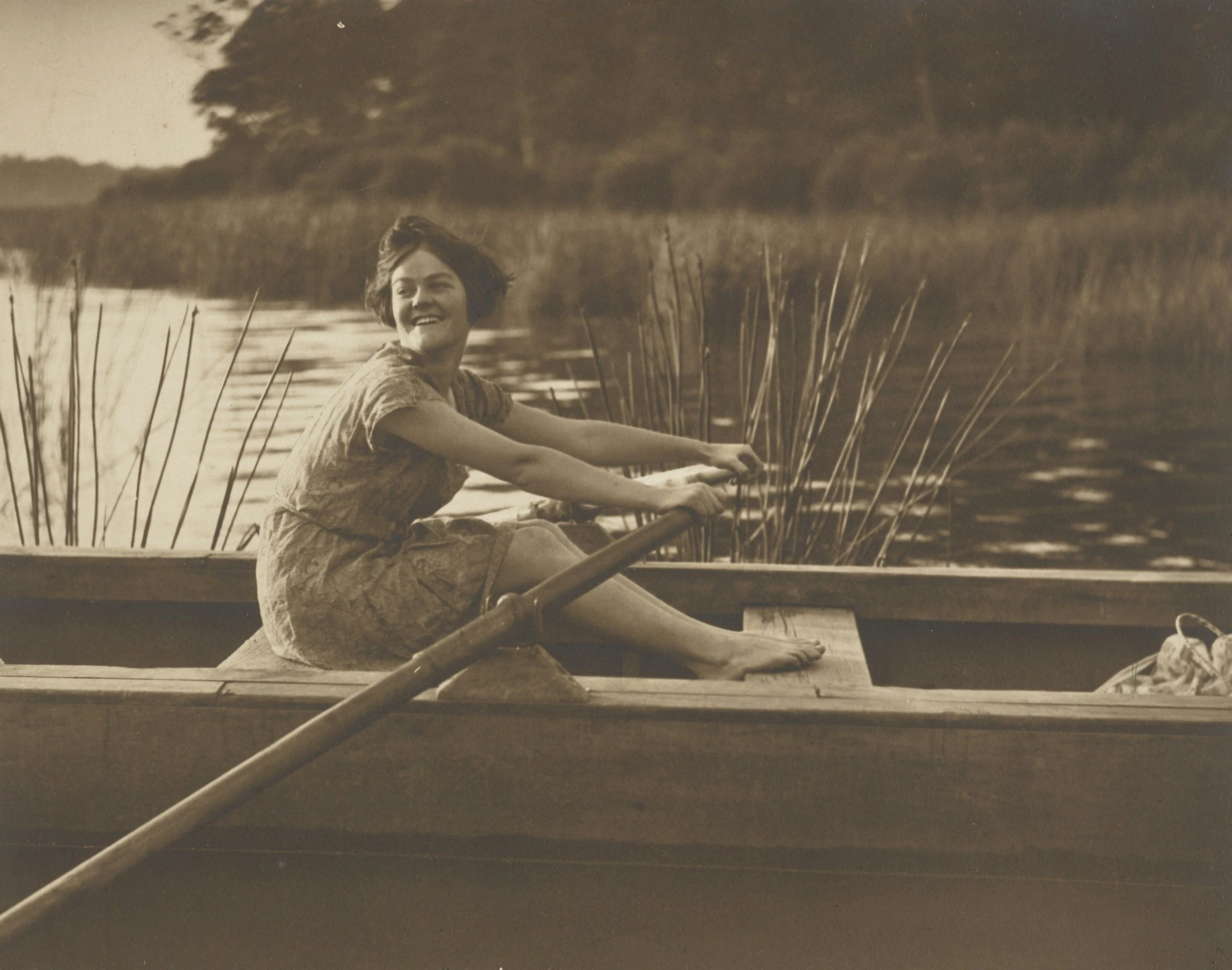 A young woman with short dark hair smiling and sitting on a boat, with a lake and trees in the background, captured in a vintage black-and-white photo.