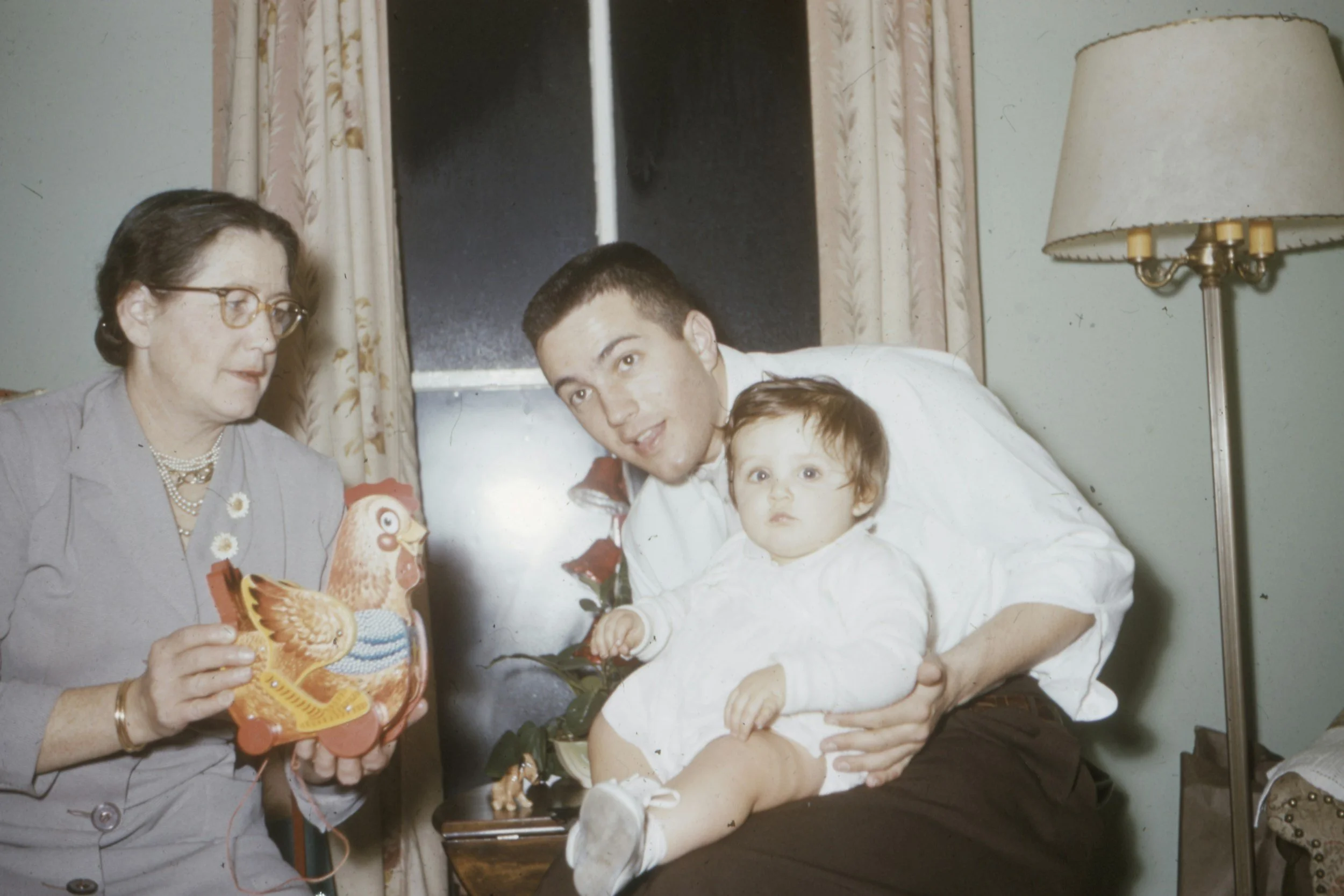 A woman in glasses and a light-colored outfit holds a colorful rooster-shaped cake, with a man holding a young girl in his arms, all smiling in a home setting with a window, curtains, and a lamp.