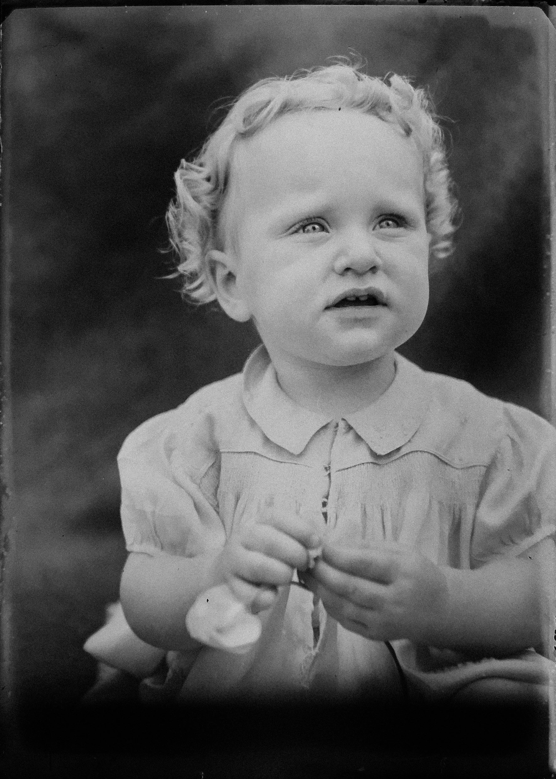 Black and white photograph of a young girl with curly hair, wearing a collared dress, looking slightly upward with a thoughtful expression, with her hands clasped together.