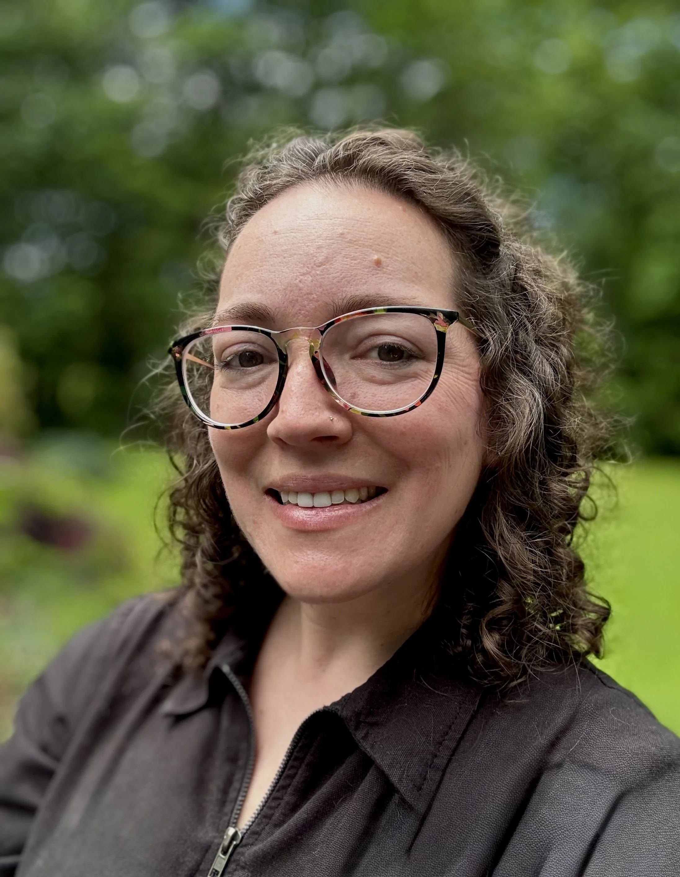 Woman with glasses smiling against a teal background, wearing a dark sweater and a white collared shirt.