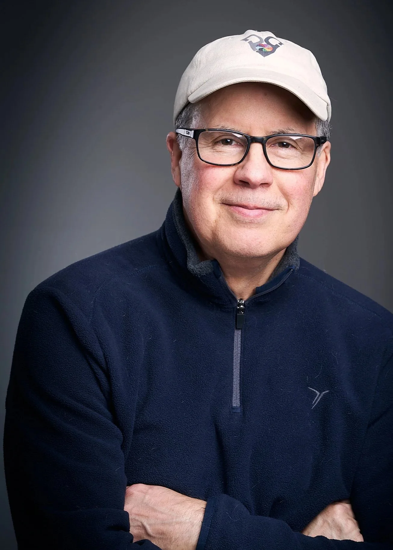 Older man wearing glasses, beige baseball cap, and dark fleece jacket, smiling against a neutral background.