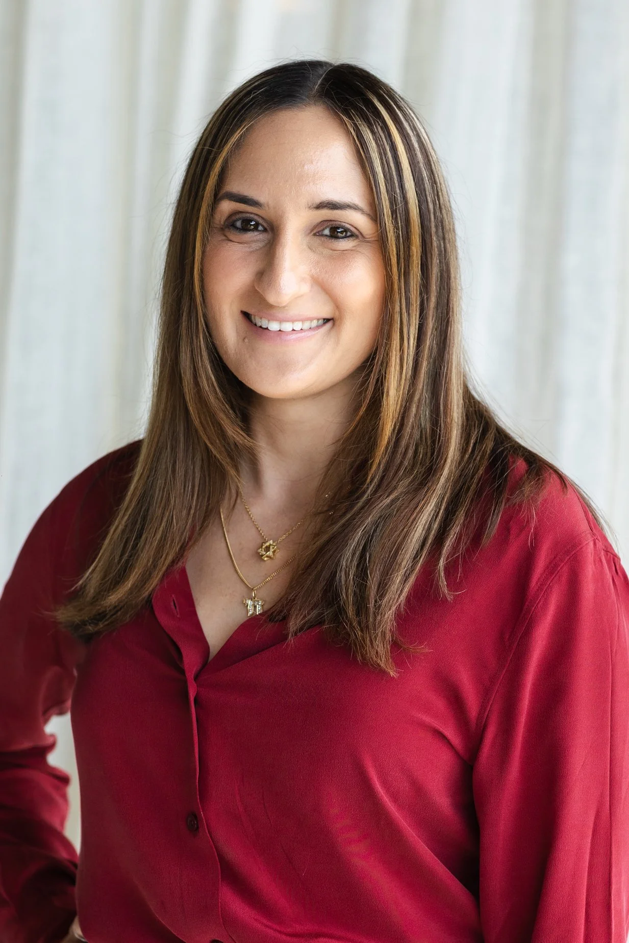 A woman with light brown hair and hazel eyes, smiling, wearing a red blouse and gold necklaces, standing indoors in front of light-colored curtains.