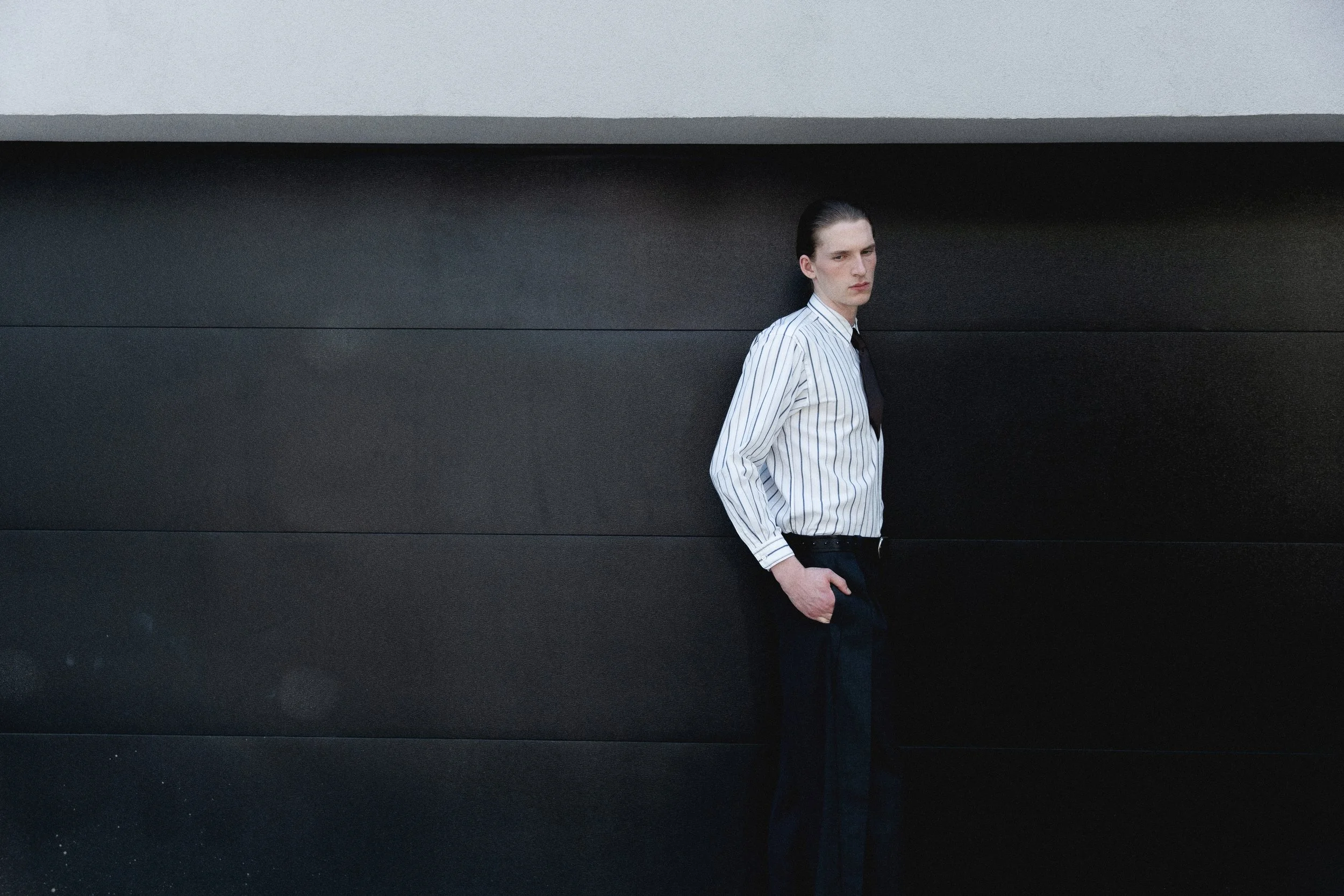 A young man with slicked-back dark hair, wearing a white striped shirt and dark pants, stands with one hand in his pocket against a black textured wall, looking slightly to the side.