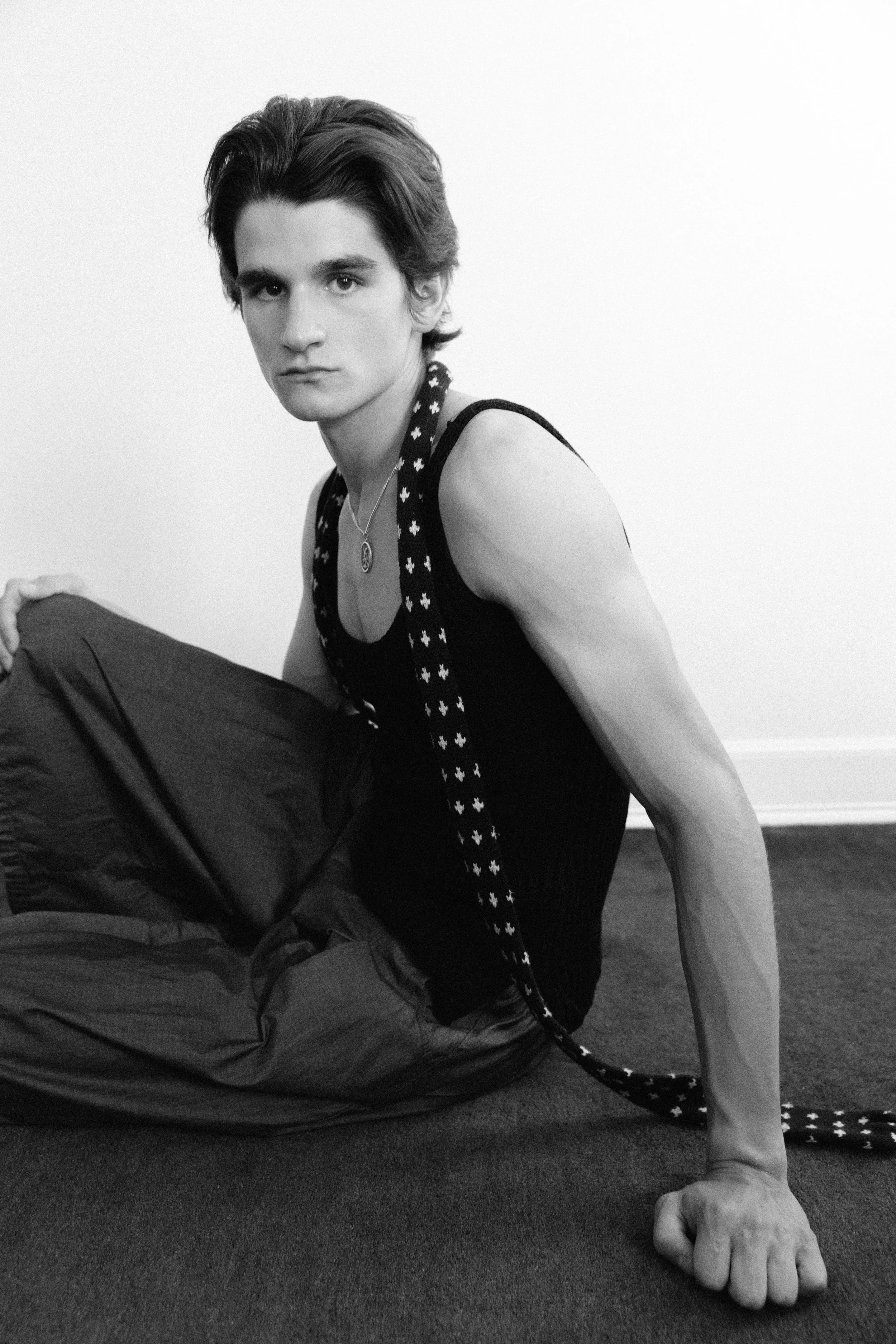 Black and white photo of a young man with dark, wavy hair, wearing a sleeveless top with a star pattern on the straps and a pendant necklace, sitting on the floor with one hand on the ground and the other on his knee, against a plain white wall backg