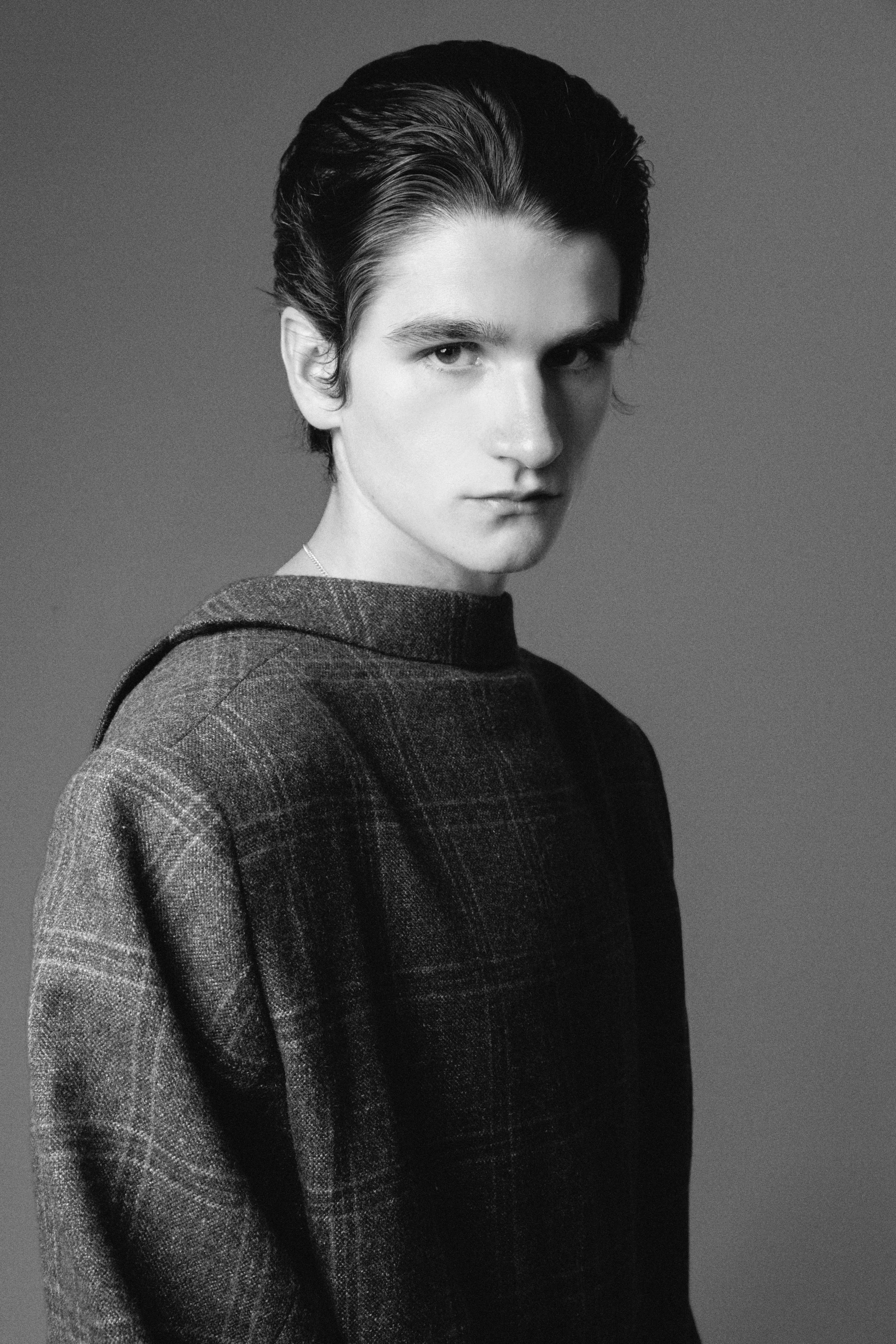 A black and white portrait of a young man with dark, slicked-back hair, wearing a plaid shirt, looking directly at the camera with a serious expression.