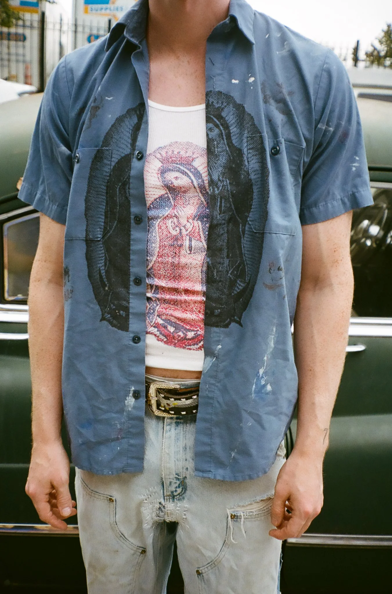 A person wearing a blue, short-sleeved button-up shirt over a white graphic t-shirt with a colorful design, beige ripped jeans, and a belt with a metallic buckle, standing in front of a vintage green car.