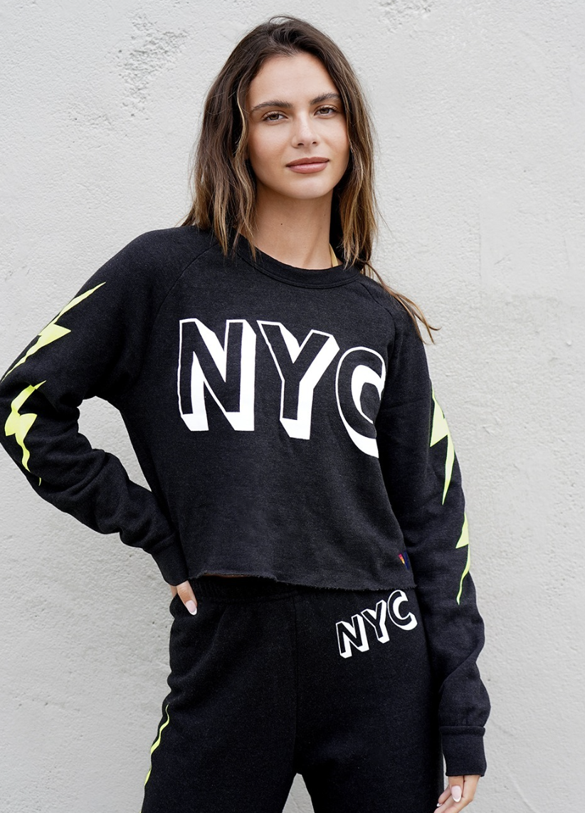 A young woman with long brown hair in a black NYC-themed sweatshirt and matching black sweatpants, standing against a light gray textured wall.