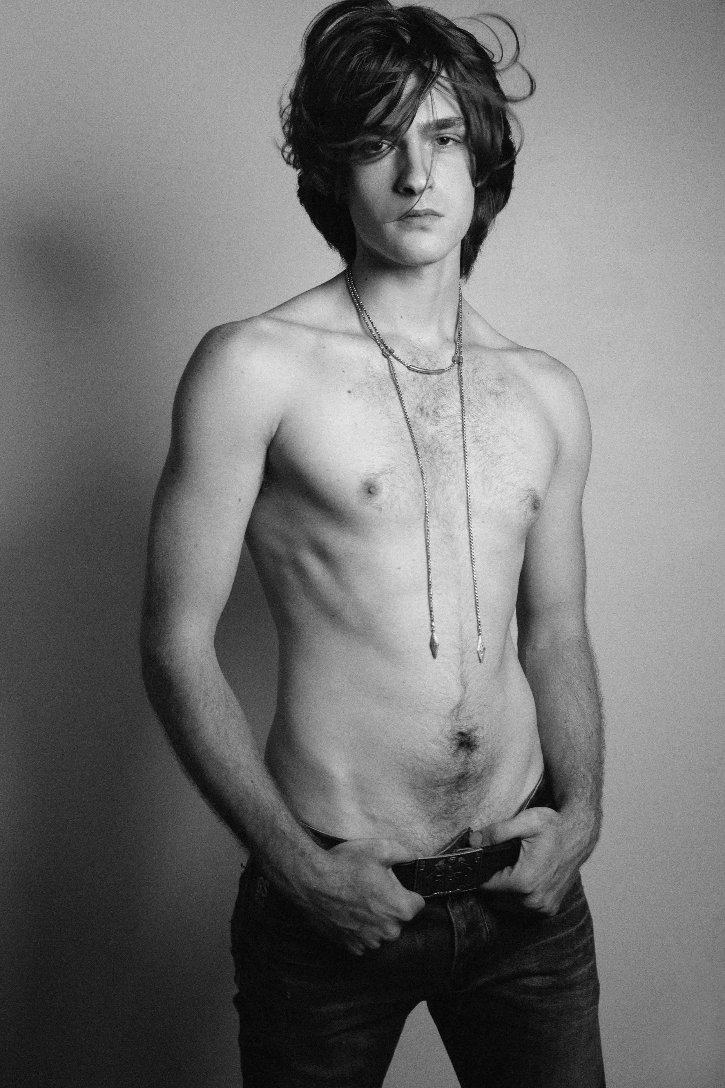 Black and white photo of a shirtless man with curly hair, wearing necklaces and jeans, standing against a plain wall.