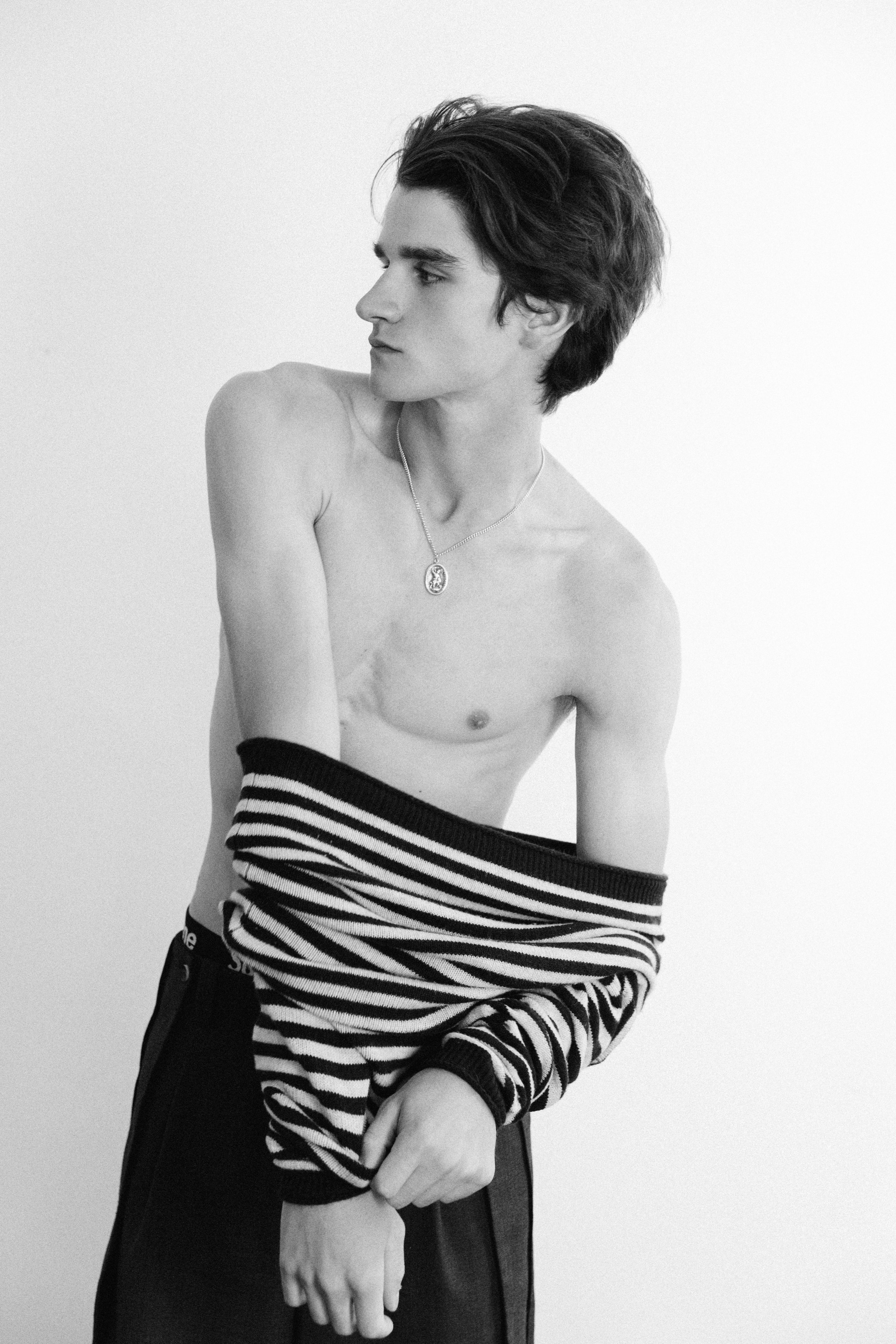 A young man with dark, tousled hair looking to his left, wearing a necklace and striped sweater around his waist, standing shirtless against a plain background.