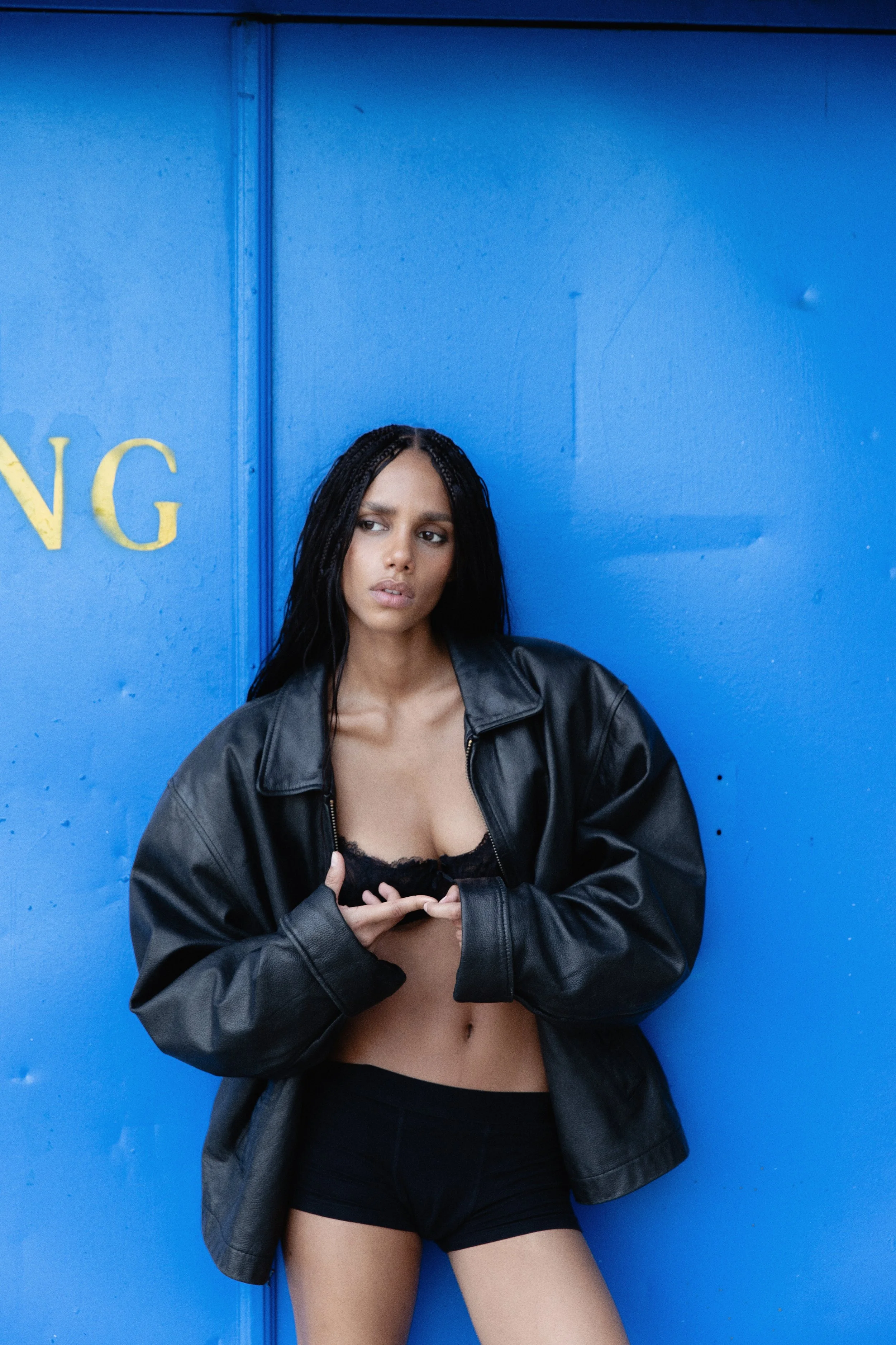 A young woman with long black hair, wearing a black leather jacket, black shorts, and a black lace bra, standing in front of a bright blue wall with partially visible yellow letters.