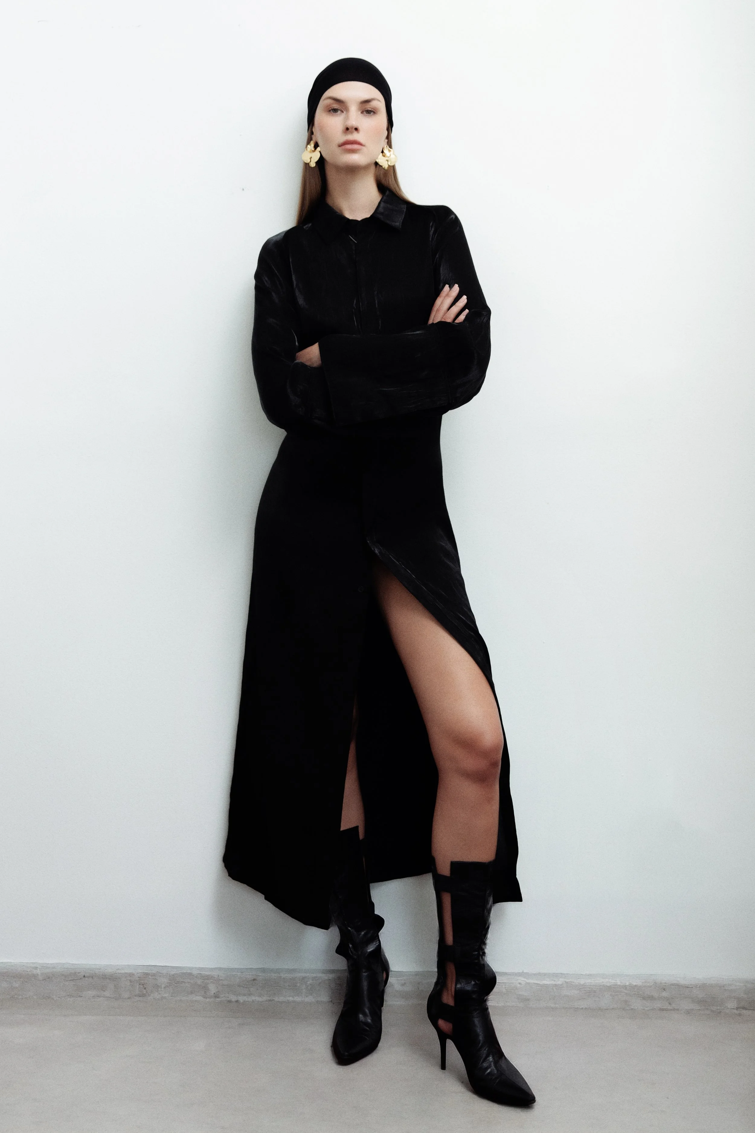 A woman standing against a white wall, wearing a black headscarf, gold earrings, a long black dress with a slit, and black high-heeled boots, crossing her arms and gazing at the camera.